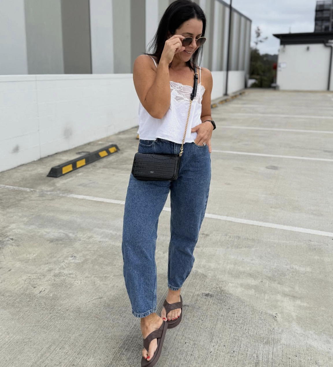 Woman wearing a white tank top, high-waisted dark wash cuffed jeans, and brown platform thong sandals while carrying a black crossbody bag.