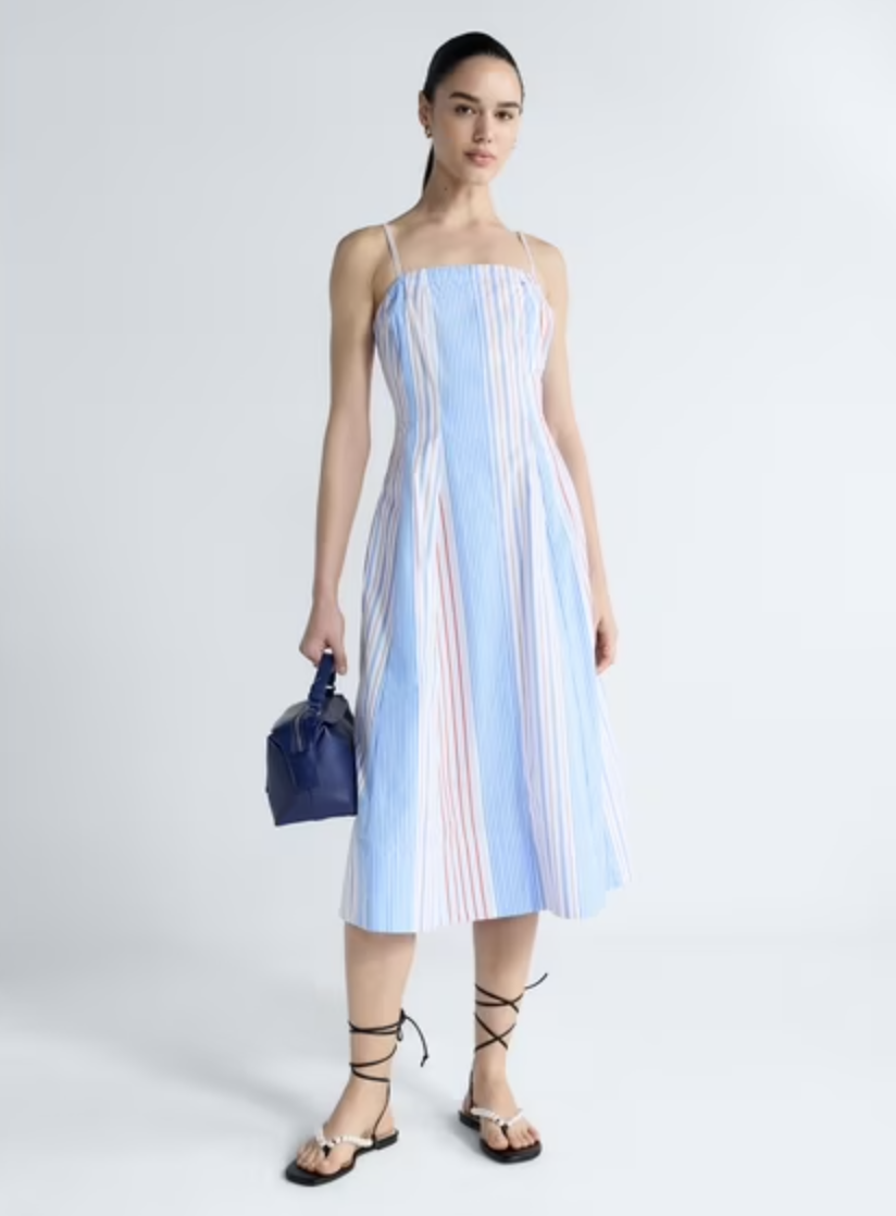 A full-length view of a woman wearing a blue and white vertical striped halter midi dress with thin red accent lines, styled with black lace-up ankle-tie sandals and a dark navy leather top-handle bag.