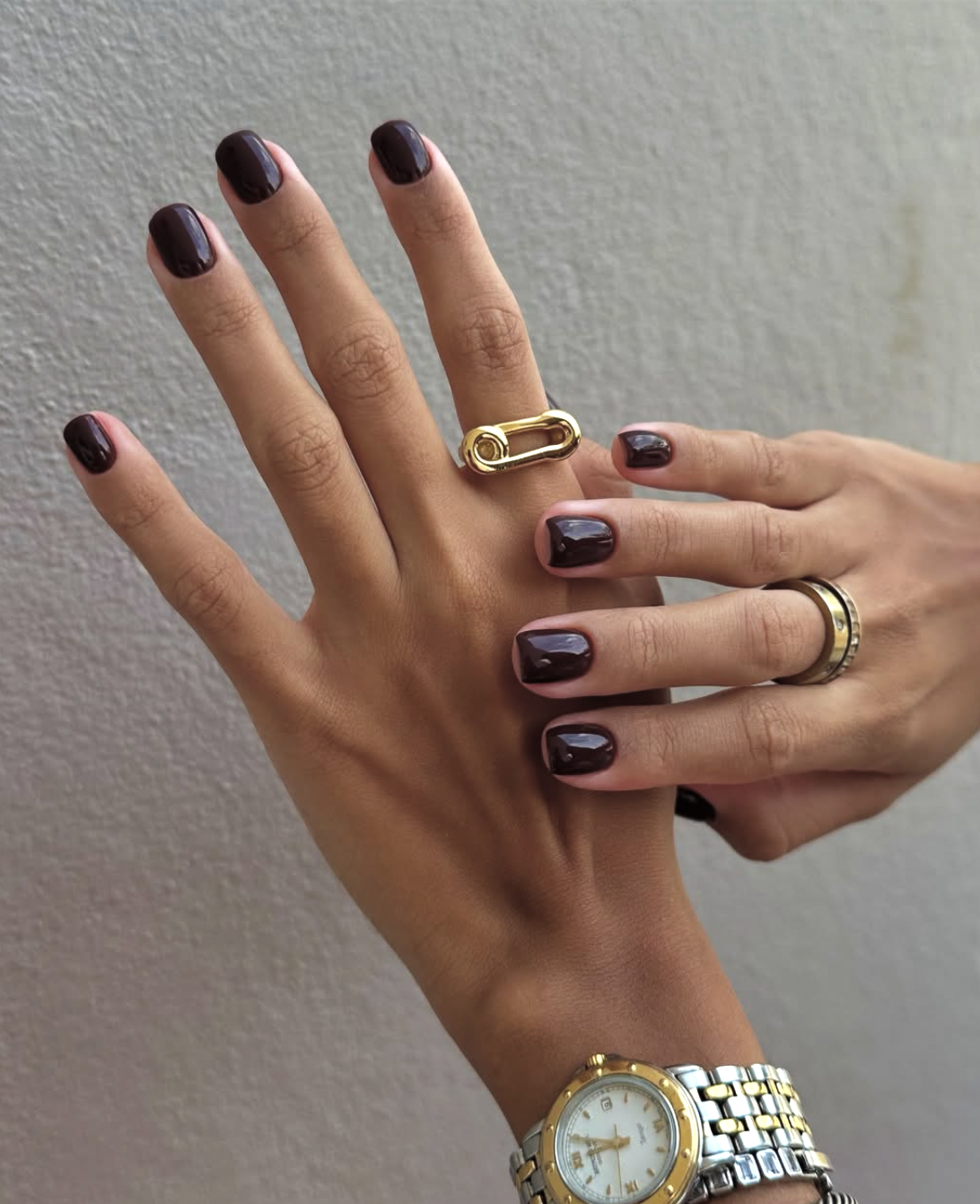 A close-up of a hand with short, squoval nails painted in a glossy, dark espresso-plum polish with gold jewelry accents.