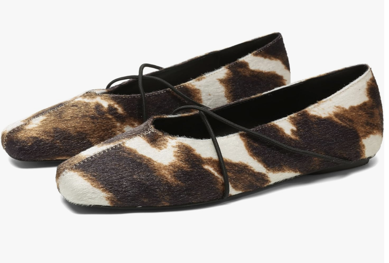 A pair of cow-print ballet flats featuring a square toe and thin black criss-cross cord straps on a white background.