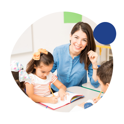 A smiling female teacher sitting at a desk with two young students, a girl and a boy, in a classroom setting. The teacher is engaging with the children as they work on a colorful activity or workbook.