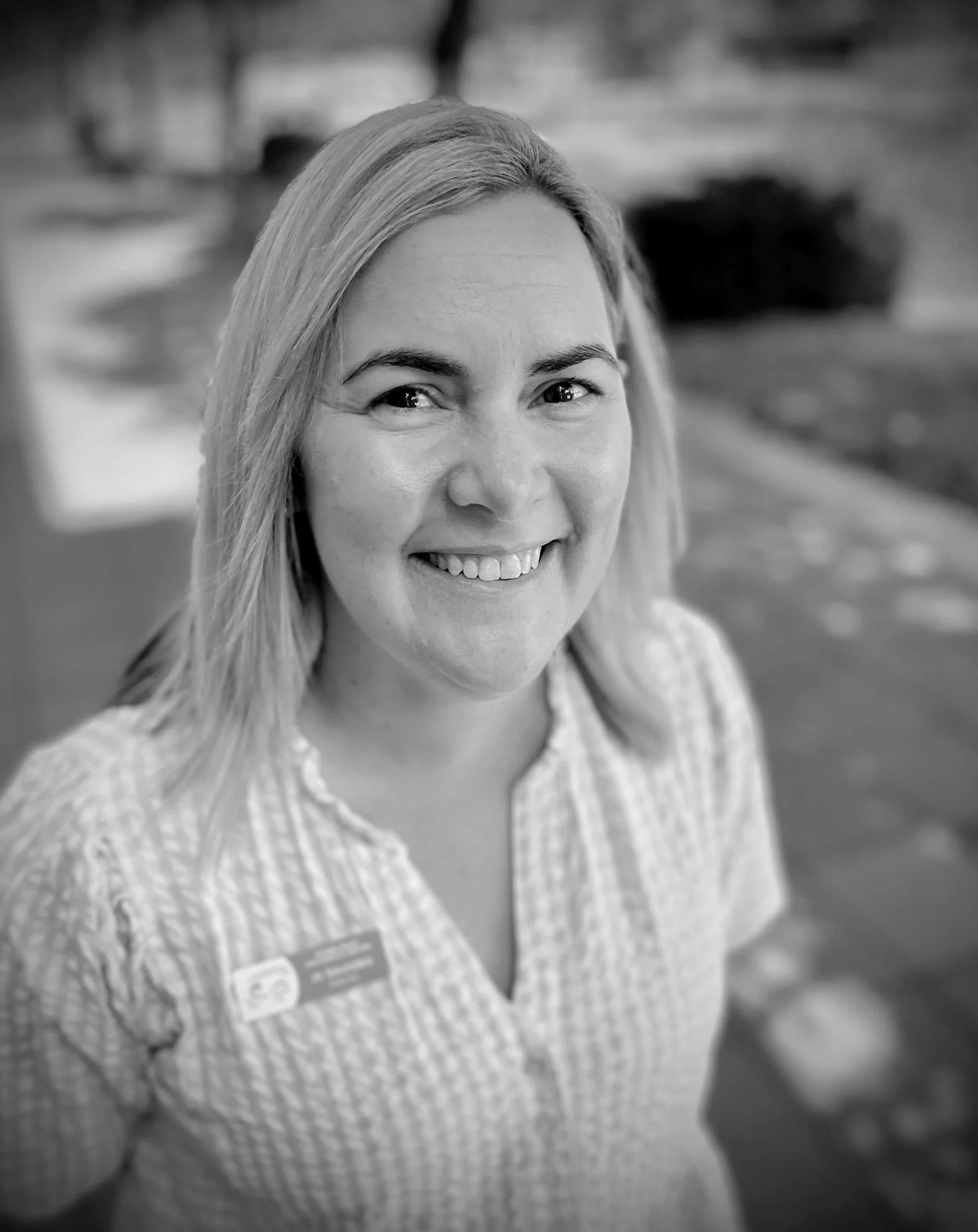 Black and white photo of a smiling woman with shoulder-length hair wearing a checkered collar shirt and a name badge.