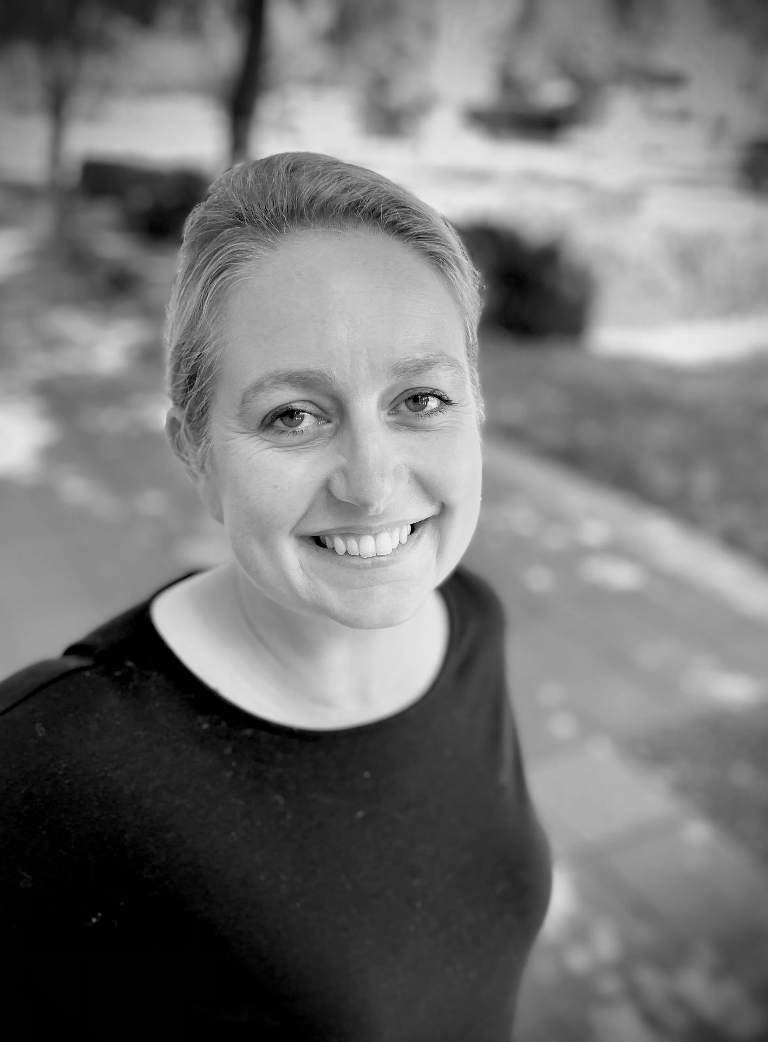 A woman with short dark hair, glasses, and earrings smiling in black and white indoors with a tree in the background.