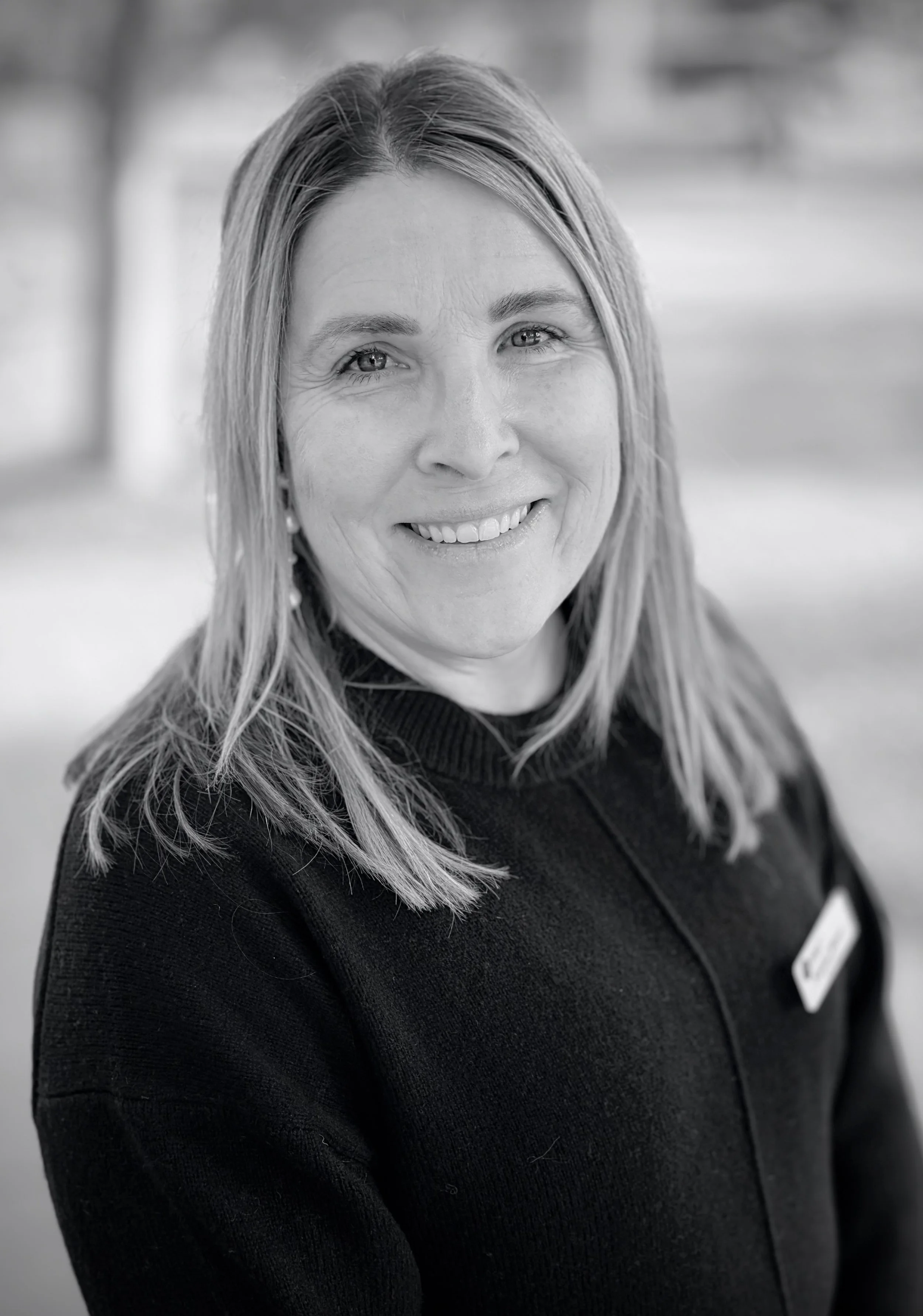 Black and white photo of a smiling woman with light-colored hair, wearing a dark top, standing outdoors with trees and a path in the background.