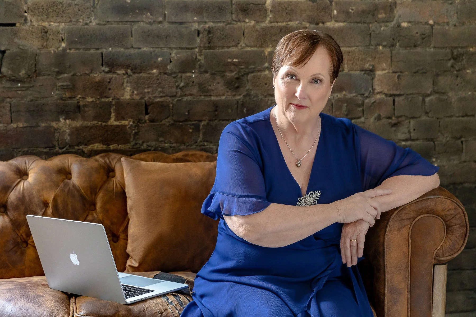 A woman in a blue dress with a laptop sitting on a brown couch.  She is the authority.