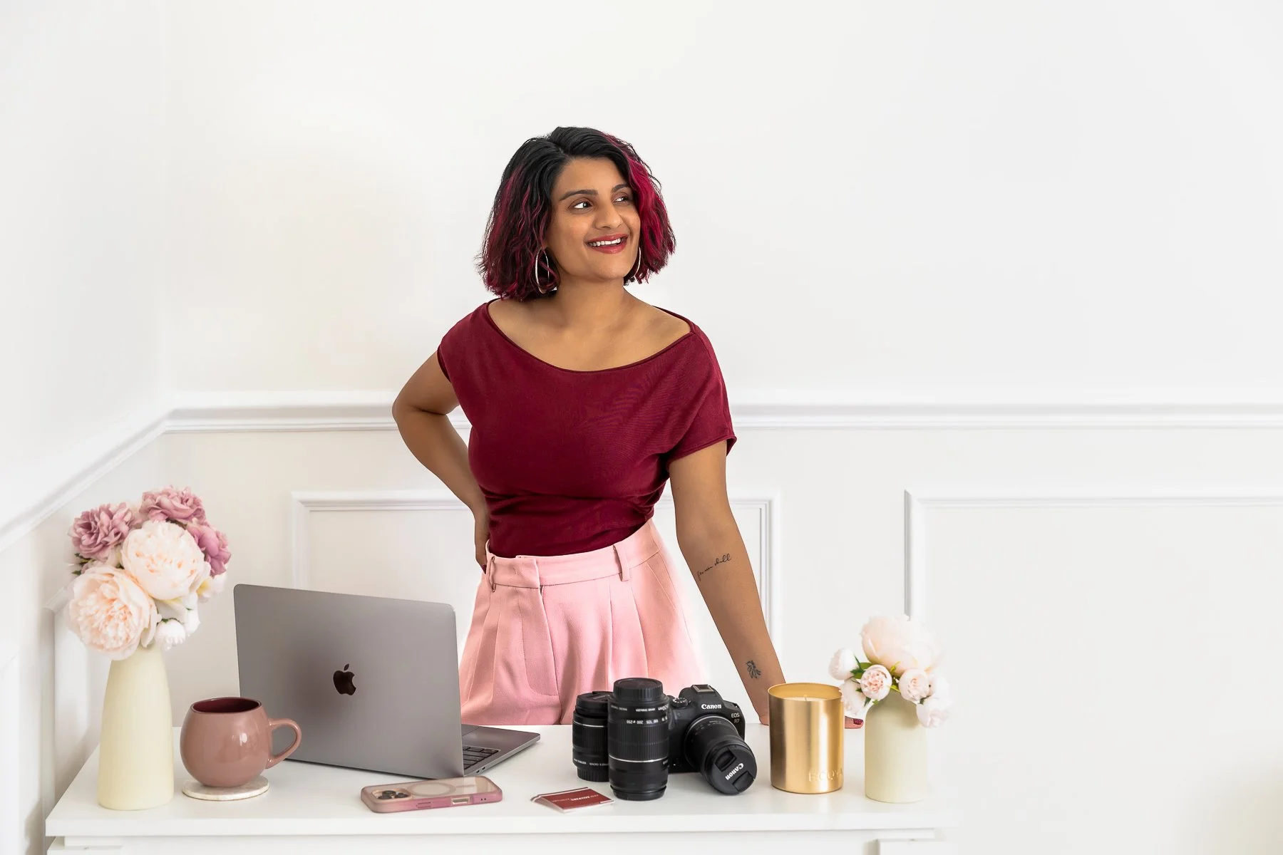 A woman dressed in pink leaning on a desk with a laptop and camera looking off to the side with a big smile.  Taken in the Castle Hill studio Sydney with a white background.