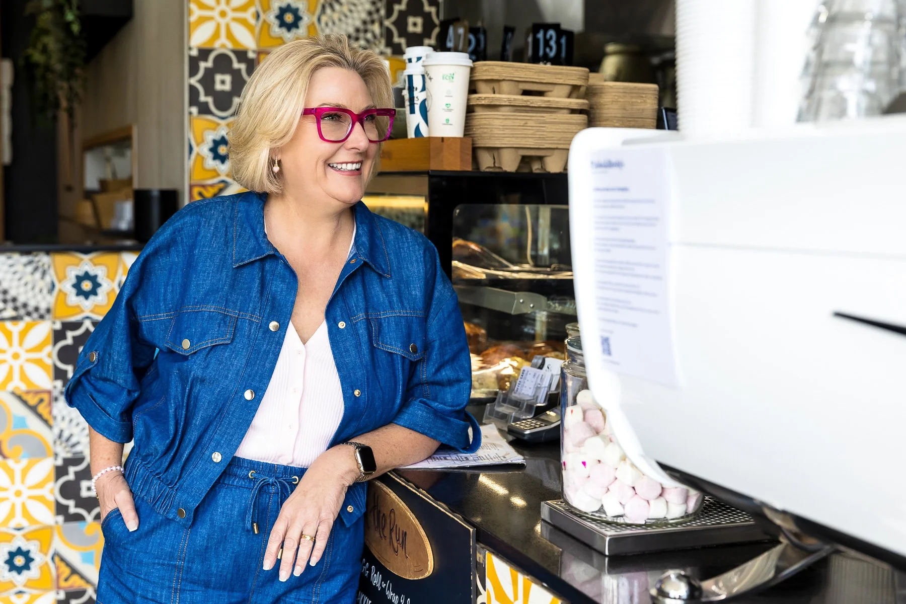 A blonde woman with pink glasses is leaning on a counter of a coffee shop and cafe in a blue outfit in the Hills District of Sydney for her personal branding shoot.
