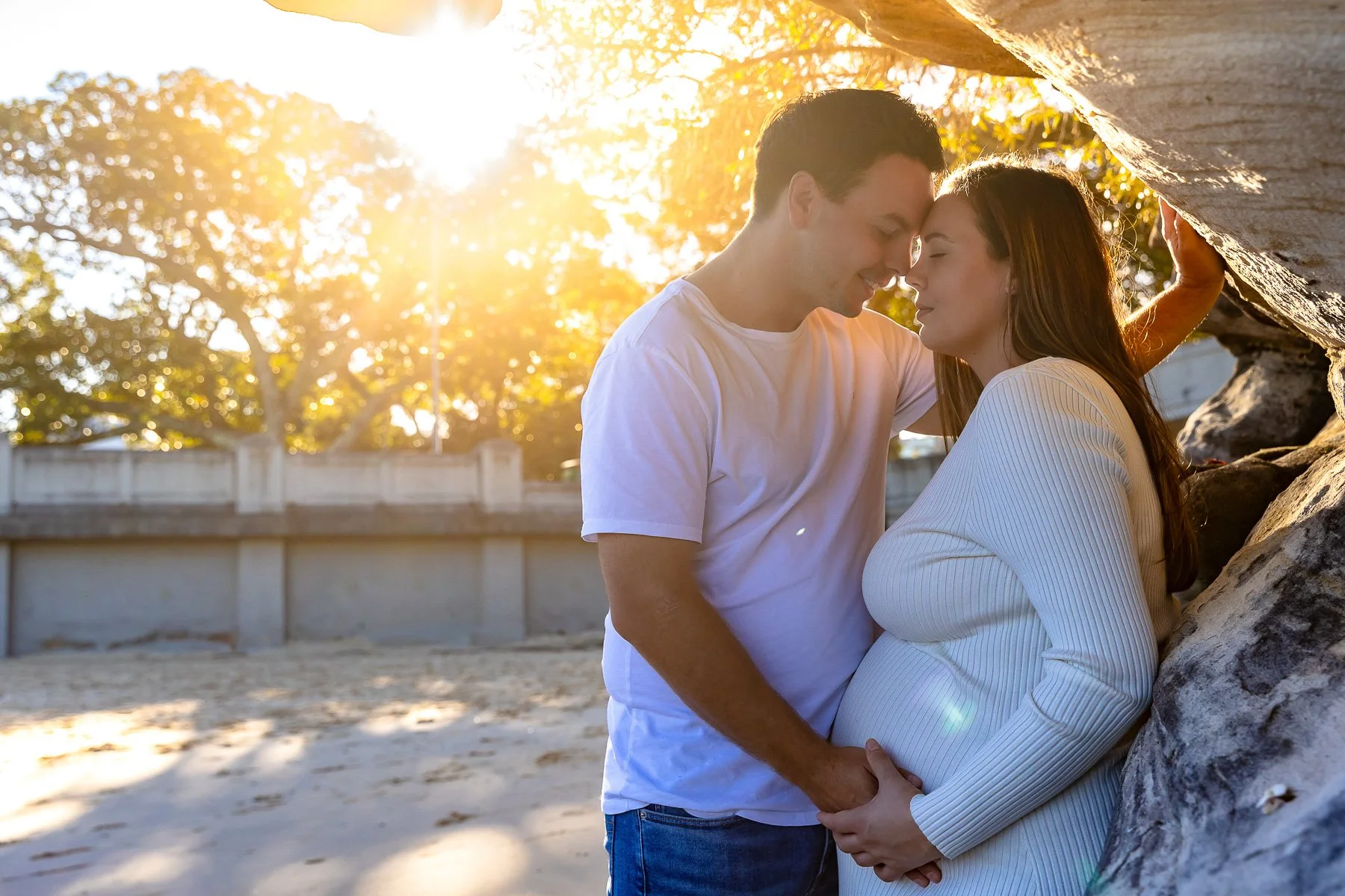 A young couple pregnant with their first child embracing in stunning afternoon sun at Balmoral Beach Sydney leaning on rocks for a maternity photoshoot