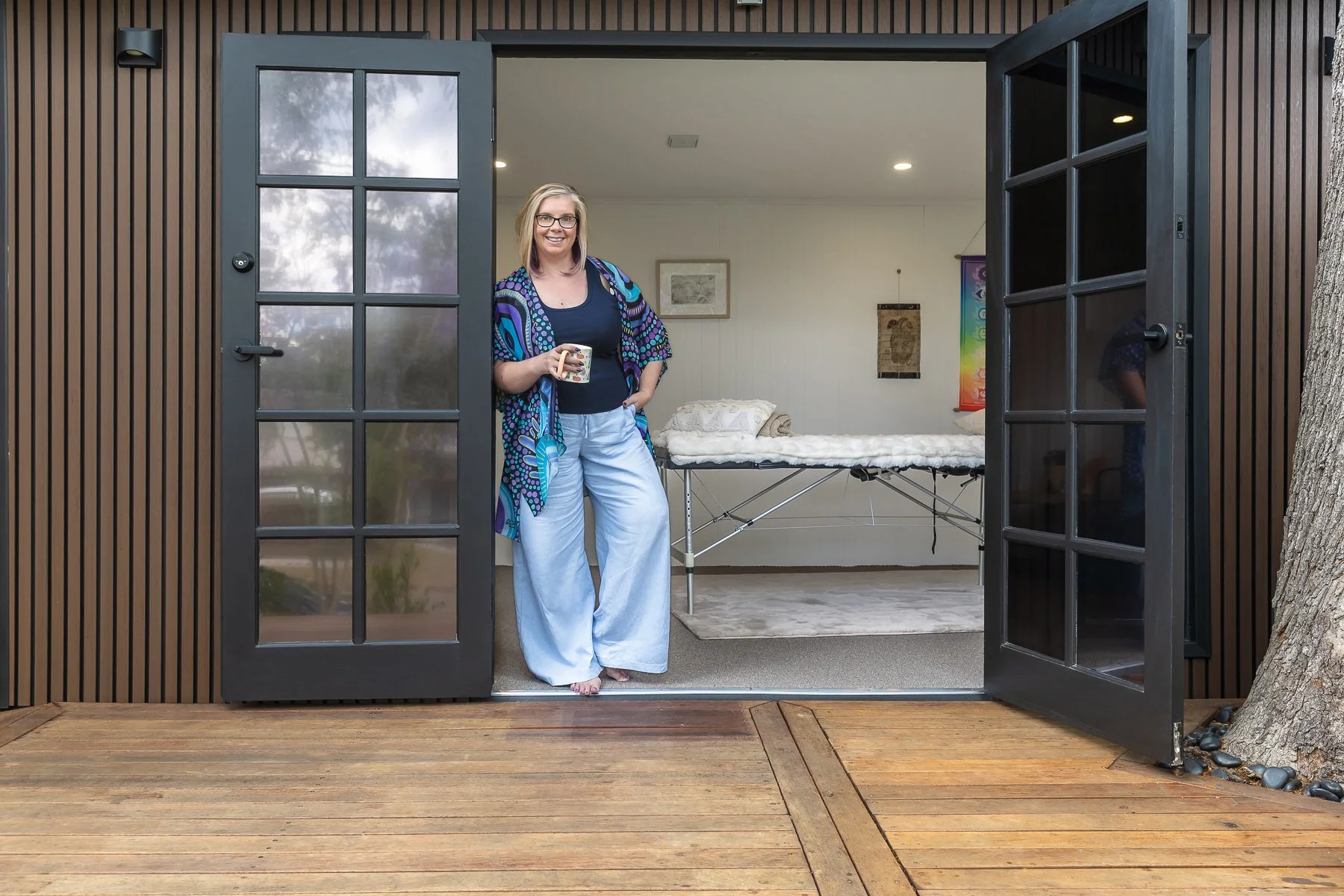 A woman is leaning on the door of her studio holding a coffee mug for her branding shoot with her massage studio in The Hills District of Sydney.