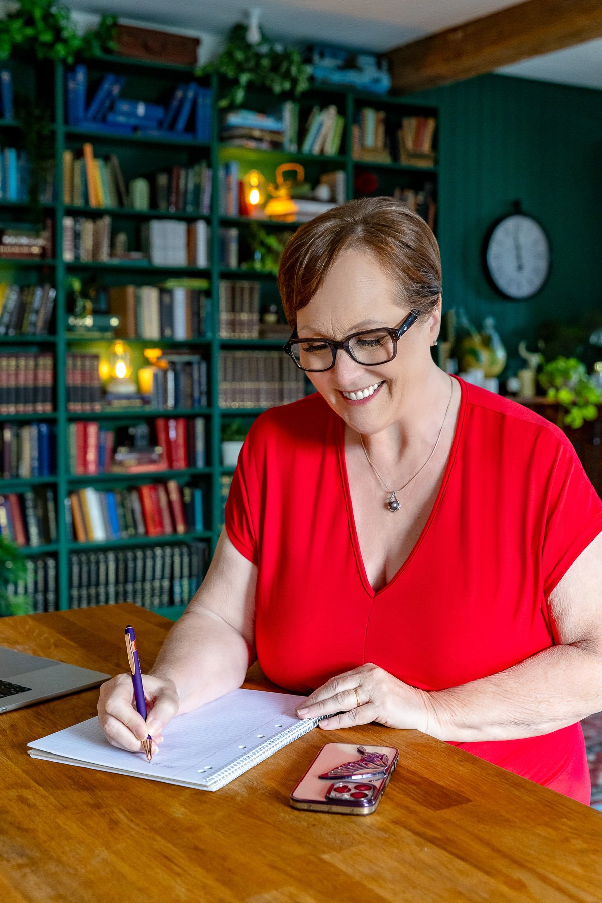 A women in a red dress with glasses is writing on her notepad with her phone and laptop next to her.  She is standing in a room with a library in the background.  Taken in a studio in Sydney.