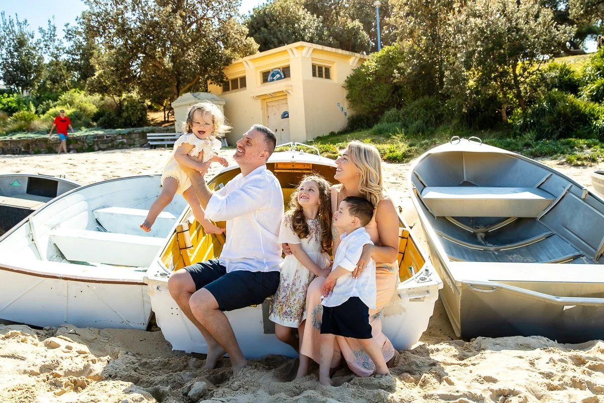 A young family sitting on the boats at Coogee Becah with dad holding the baby up in the air during a morning shoot at Coogee Beach.