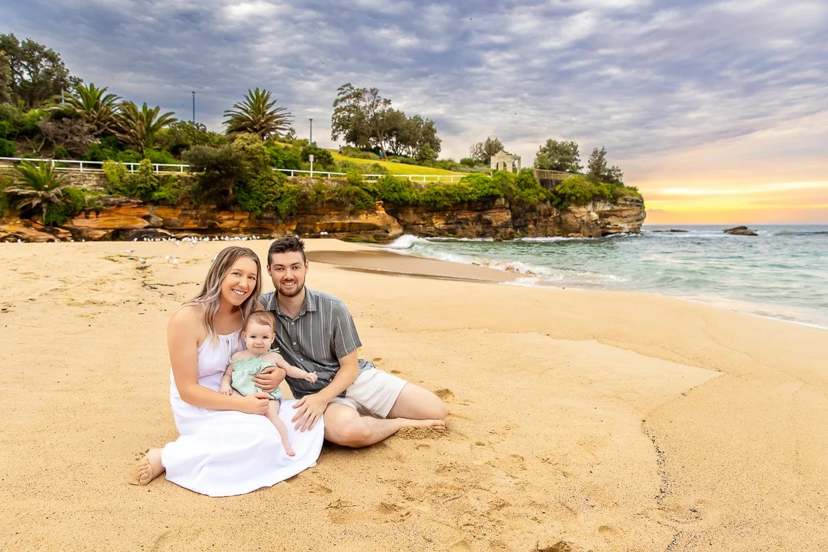 A young family with a baby sitting on the sand at Coogee Beach in Sydney's Eastern Suburbs with a gorgeous muted sunrise behind them 