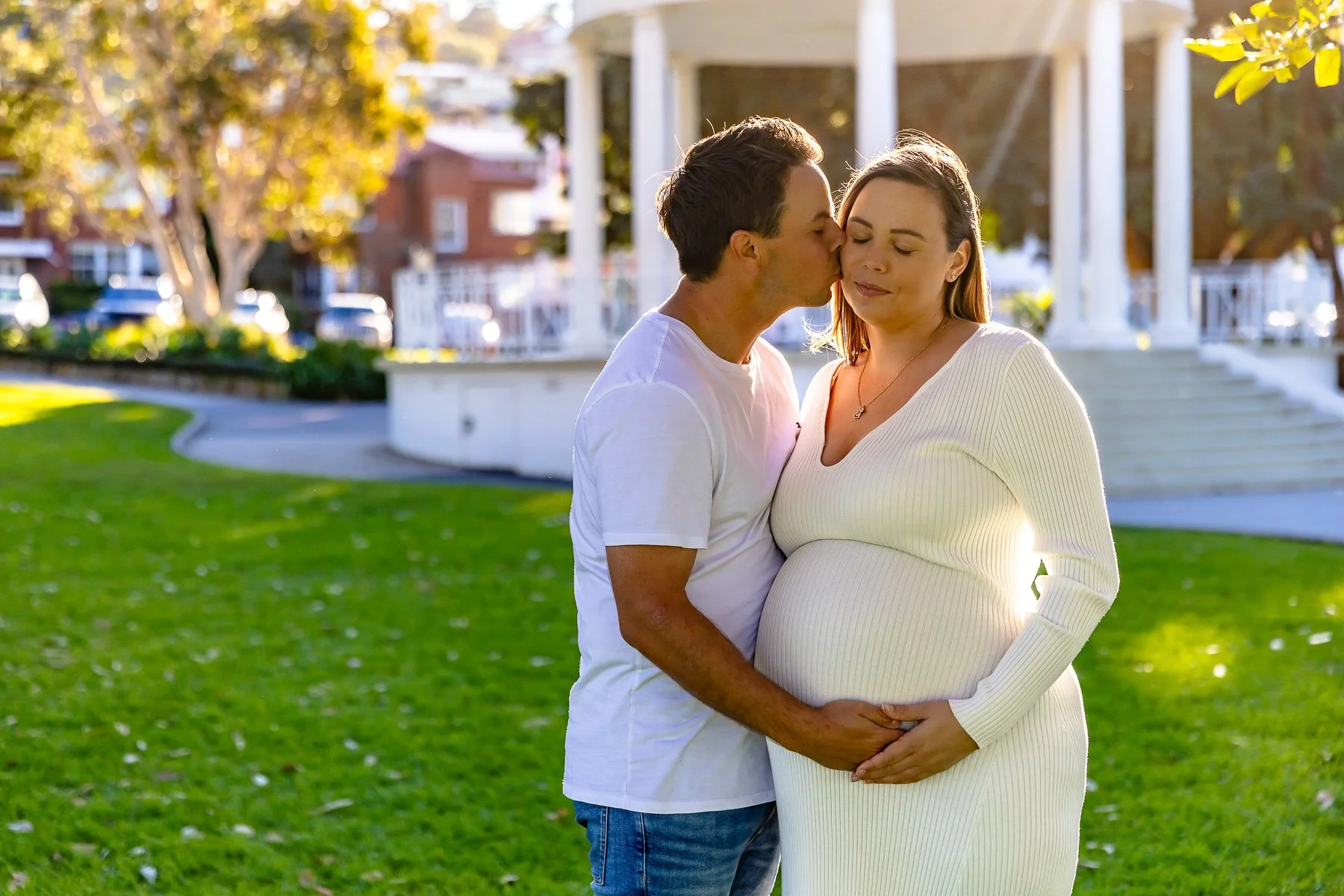 A young couple kissing in the afternoon sun with the Balmoral Beach Rotunda in the background during a maternity photoshoot
