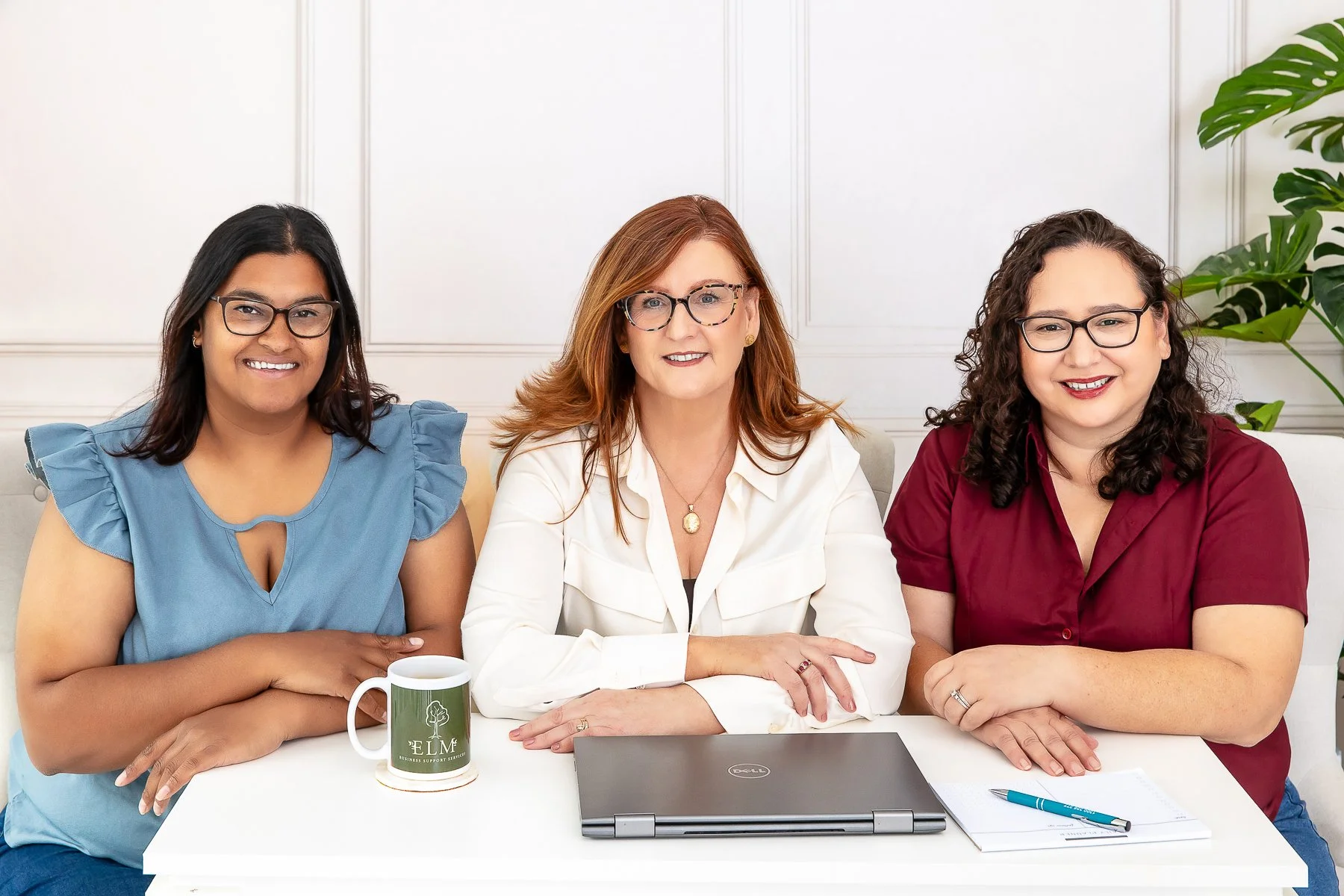 Three women sitting at a desk with a laptop, mug, notepad and pen.