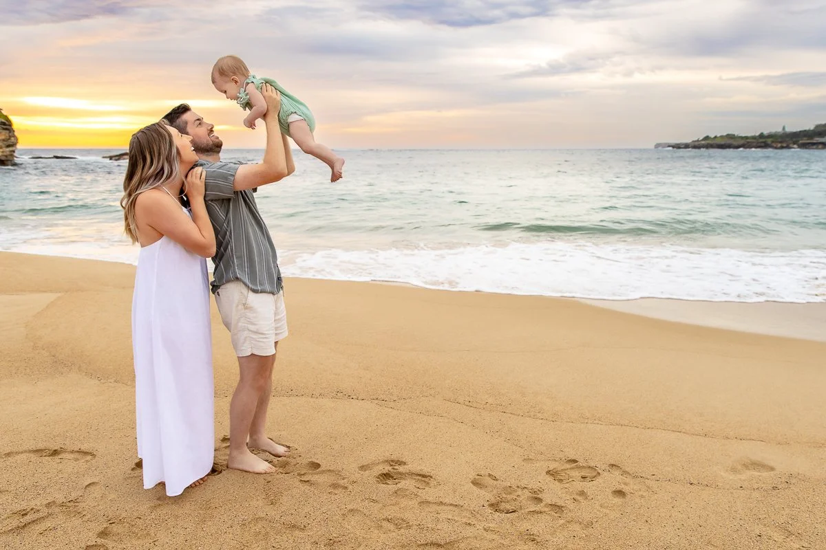 A young family holding their baby girl up in the air on Coogee Beach with muted sunrise colours behind them