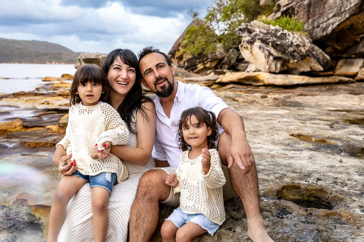 A gorgeous young family sitting on the rocks at Balmoral Beach in Sydney embracing and enjoying the afternoon together during a family photoshoot.