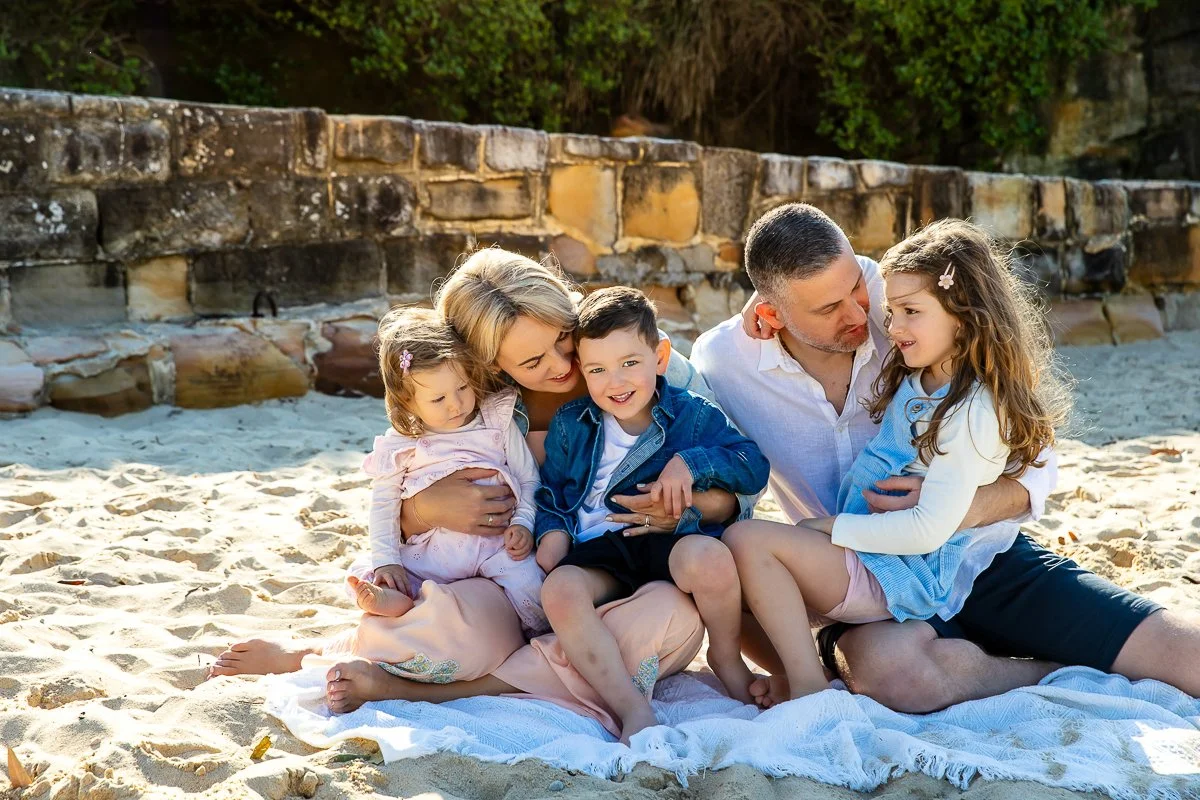A young family cuddling on a blanket on the sand at Coogee Beach in the morning light for their outdoor family photoshoot