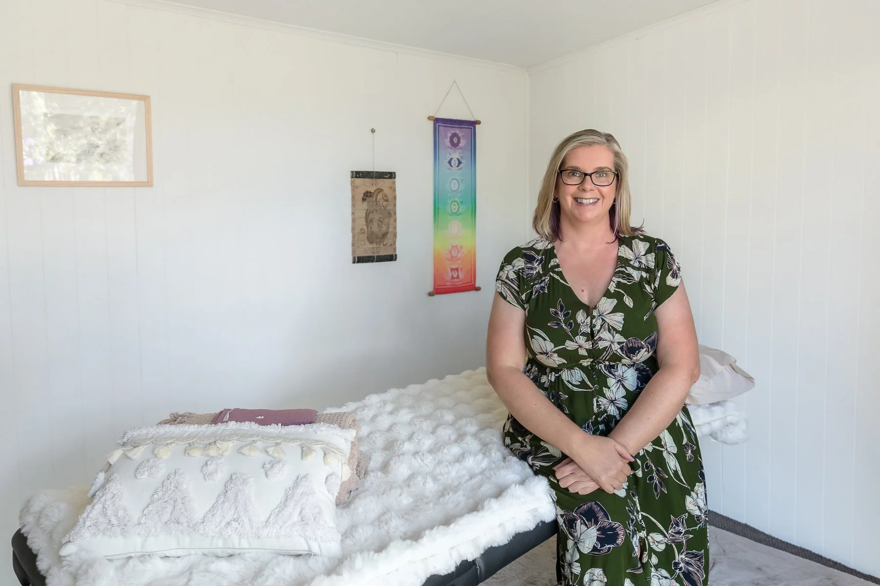 A woman sitting on a massage table with pillow and wall hangings behind her showcasing her studio for her branding shoot