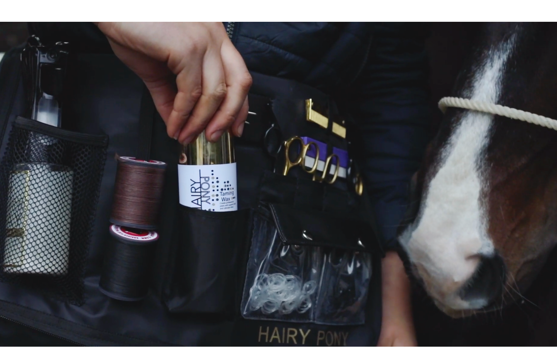 Close-up of a person's hand grooming a horse using tack and grooming supplies on a black grooming bag labeled 'HAIRY PONY', with grooming tools, thread spools, and a container of hair ties visible, while the horse's head is partially visible.