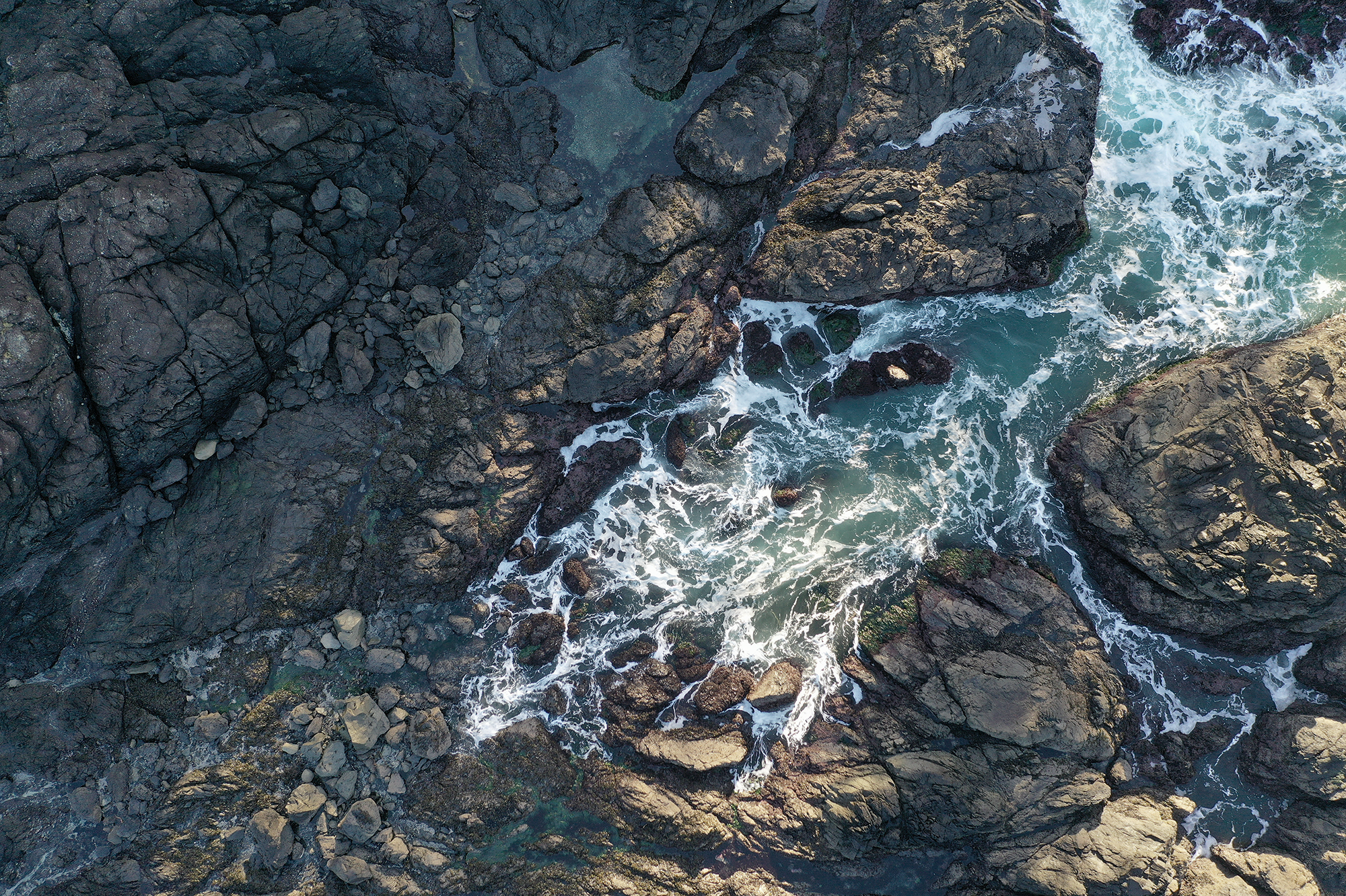 Aerial view of a rocky shoreline with ocean waters crashing against the rocks and swirling tide pools.