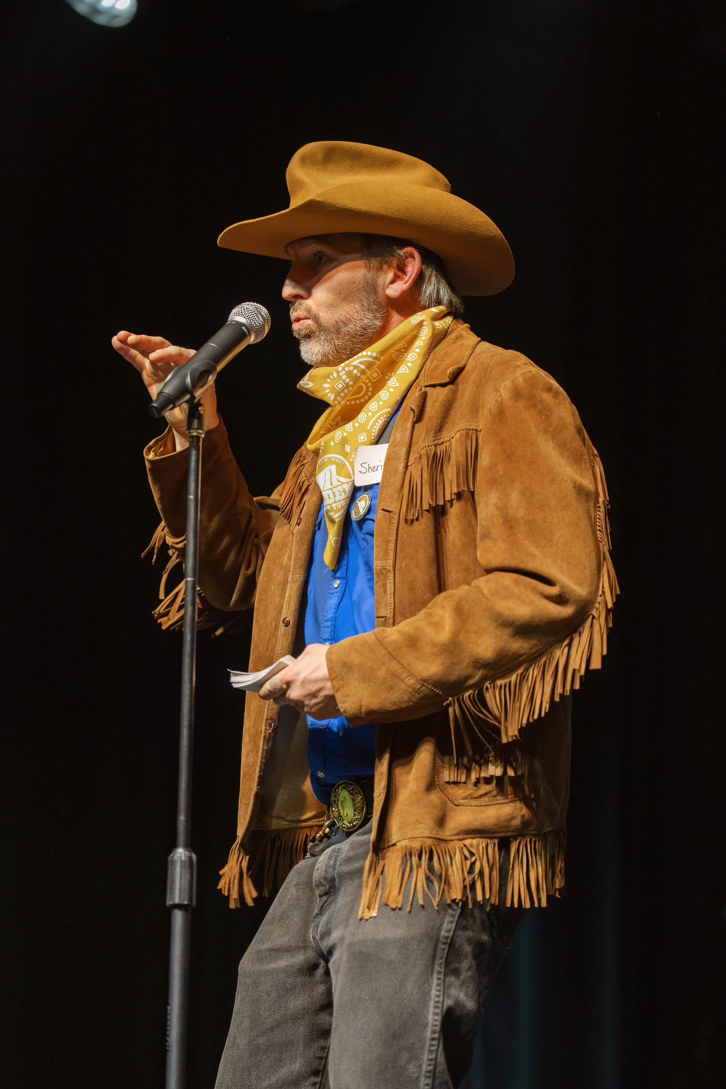 A man dressed in western attire talking into a microphone on a stage.