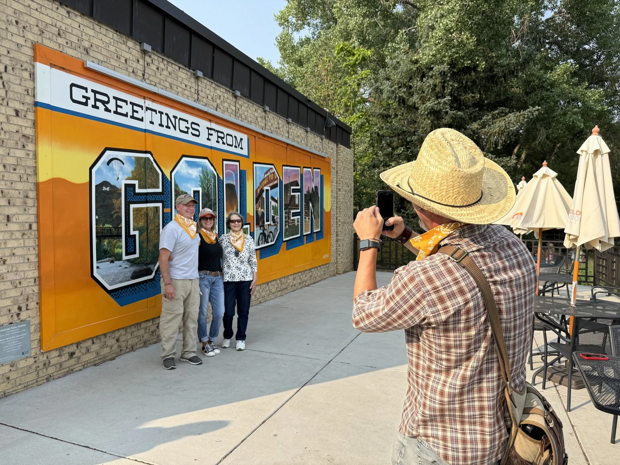 A man in western wear takes a photo of a group of people in front of a mural.
