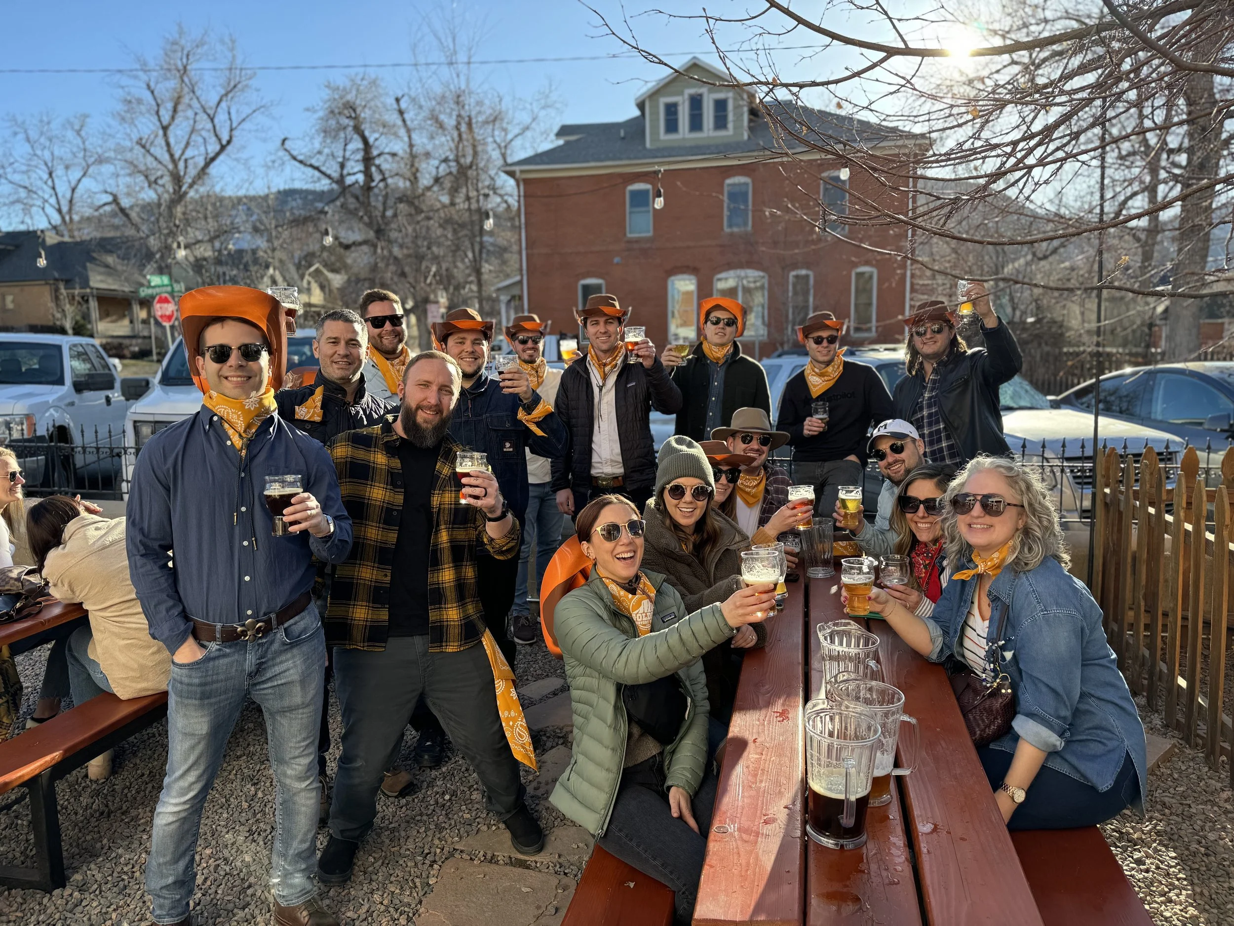 People standing and sitting around picnic tables dressed in western attire holding up their beer glasses.
