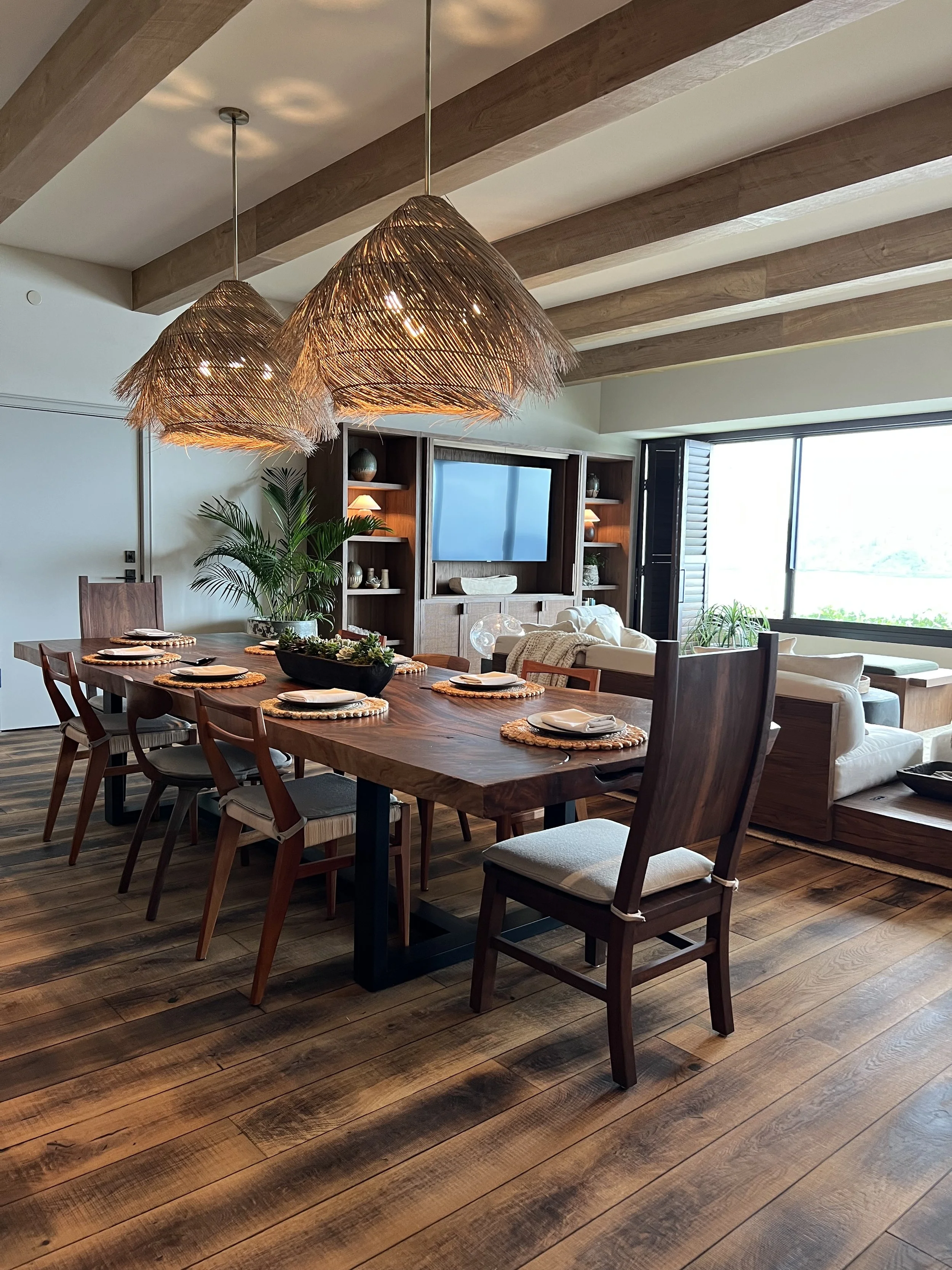 Dining room with a large wooden table set with plates and placemats, surrounded by wooden chairs. Overhead, three woven pendant lights hang. In the background, a cozy living area with a sofa, a large window, and a TV unit.