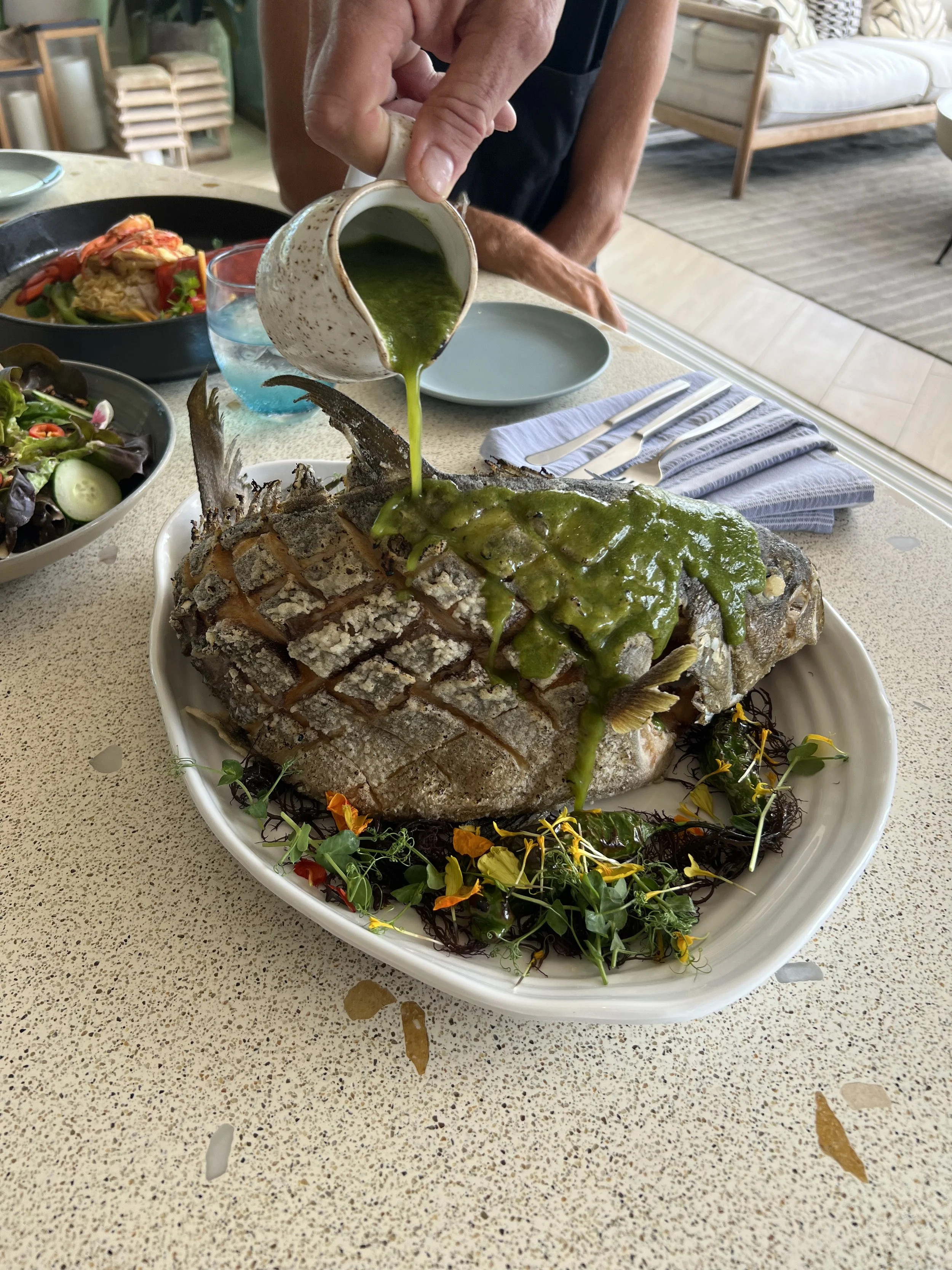 A cooked fish on a platter, being topped with green sauce poured from a small pitcher, garnished with edible flowers and herbs, with side salads and utensils on the table.