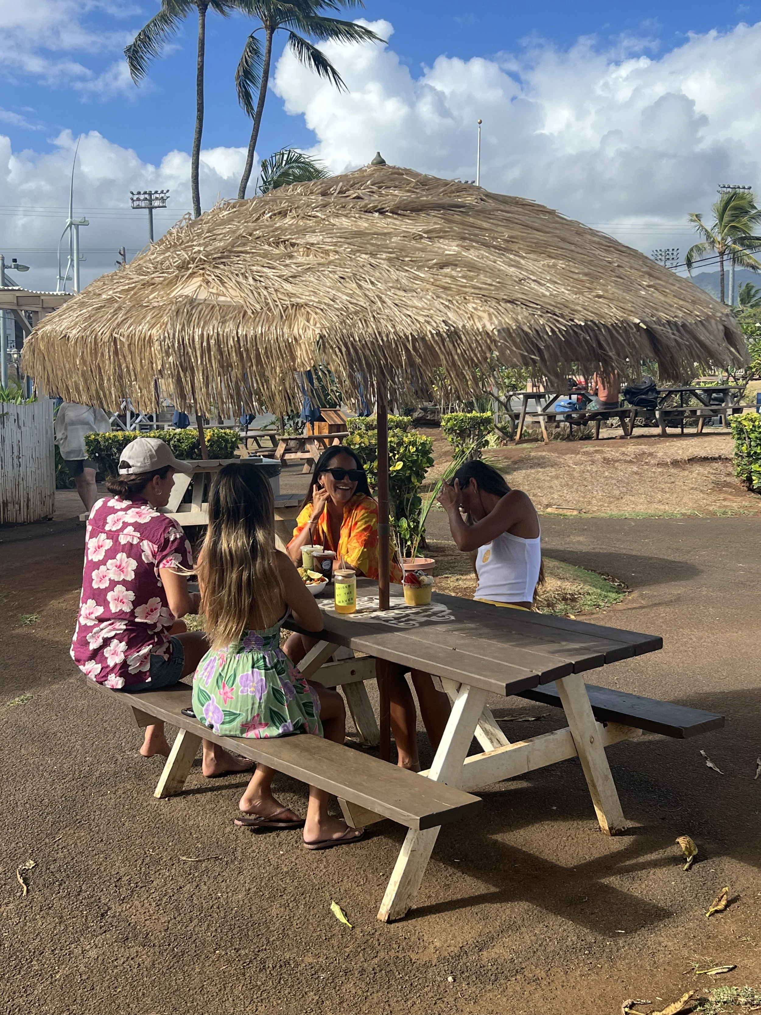 Four women sitting at a picnic table under a thatched umbrella, enjoying drinks and conversation on a sunny day.