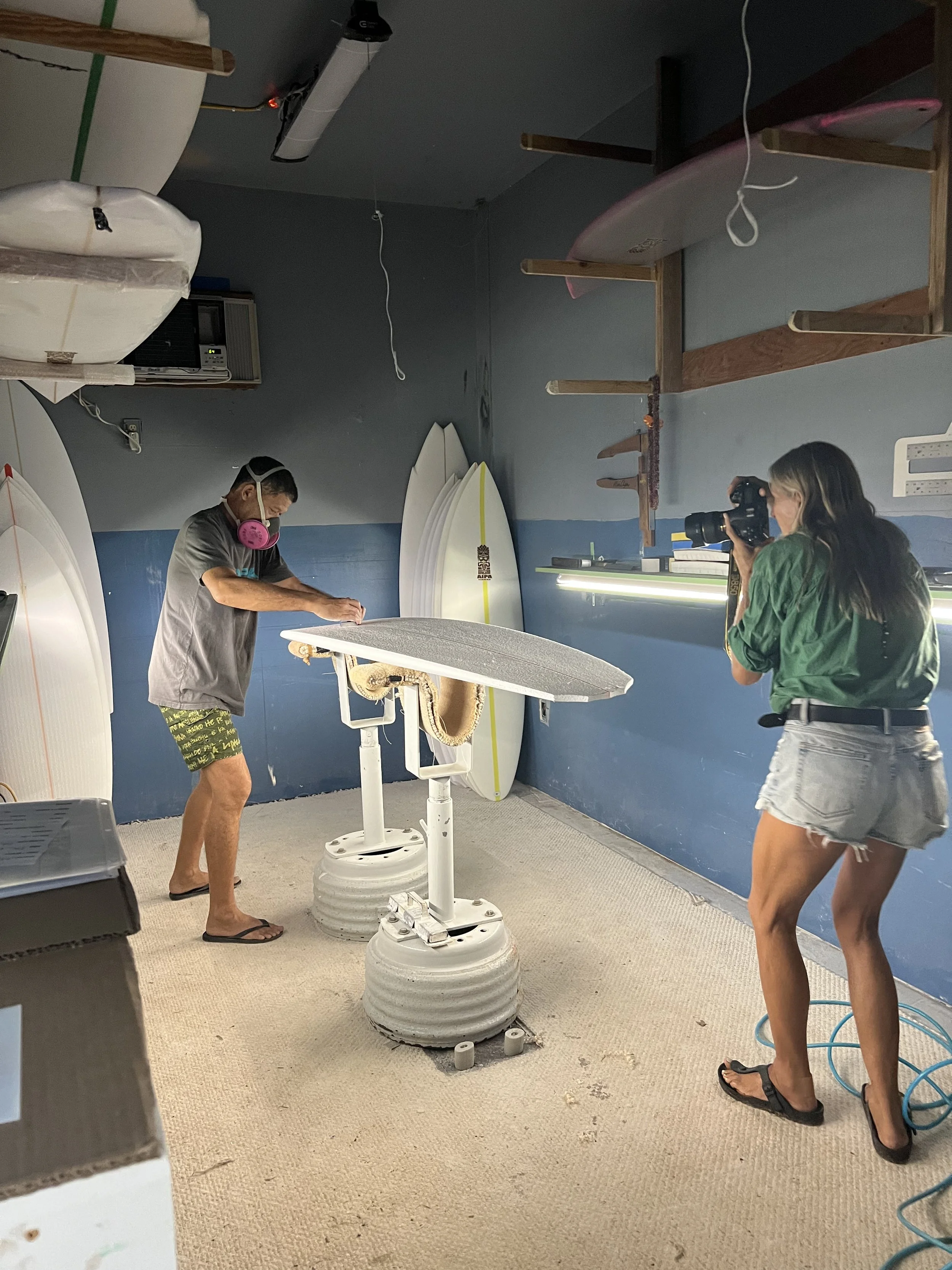 A man polishing a surfboard on a padded table while a woman takes photos in a surfboard shop or workshop with surfboards against the wall and surfboard racks above.
