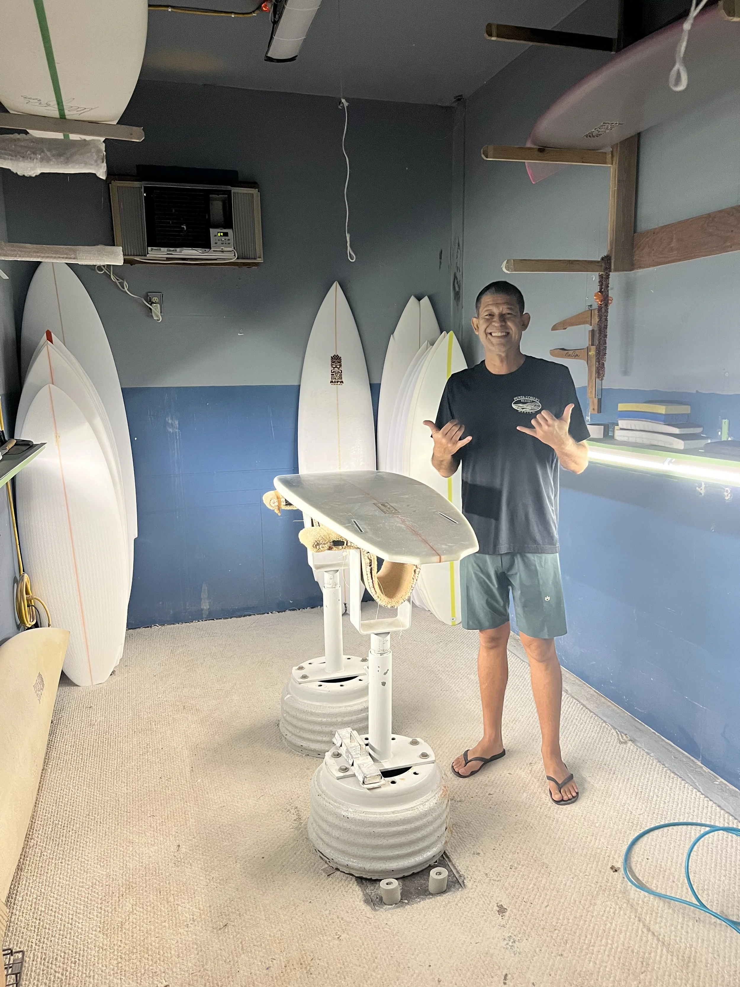 A man smiling and making a shaka sign with both hands stands next to surfboards and a stand-up paddleboard in a surf shop or repair shop, with surf-related equipment and shelves in the background.