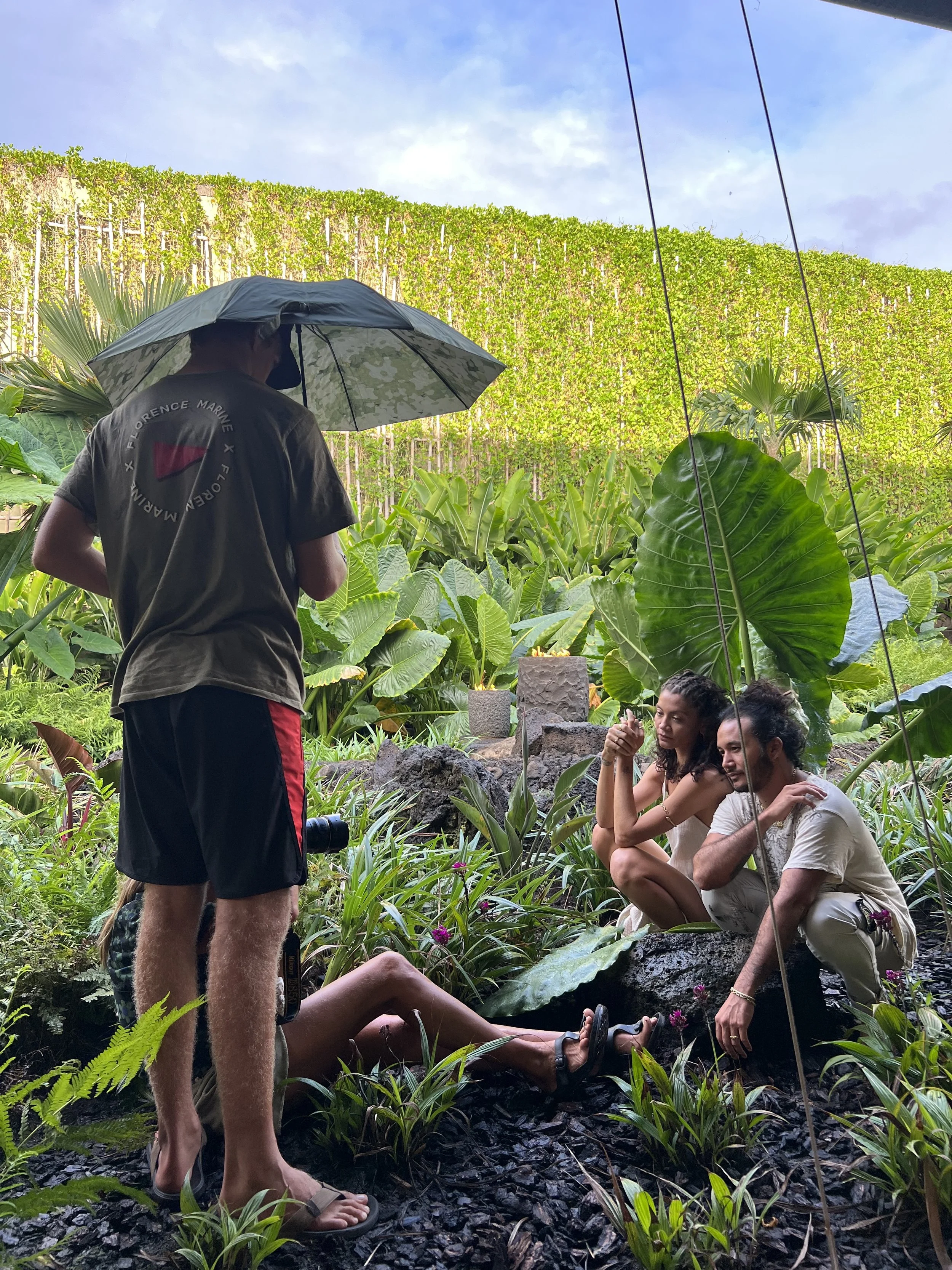 A photographer taking pictures of two people sitting on rocks in a lush jungle setting, with a person standing nearby holding an umbrella to shield from rain or sunlight.