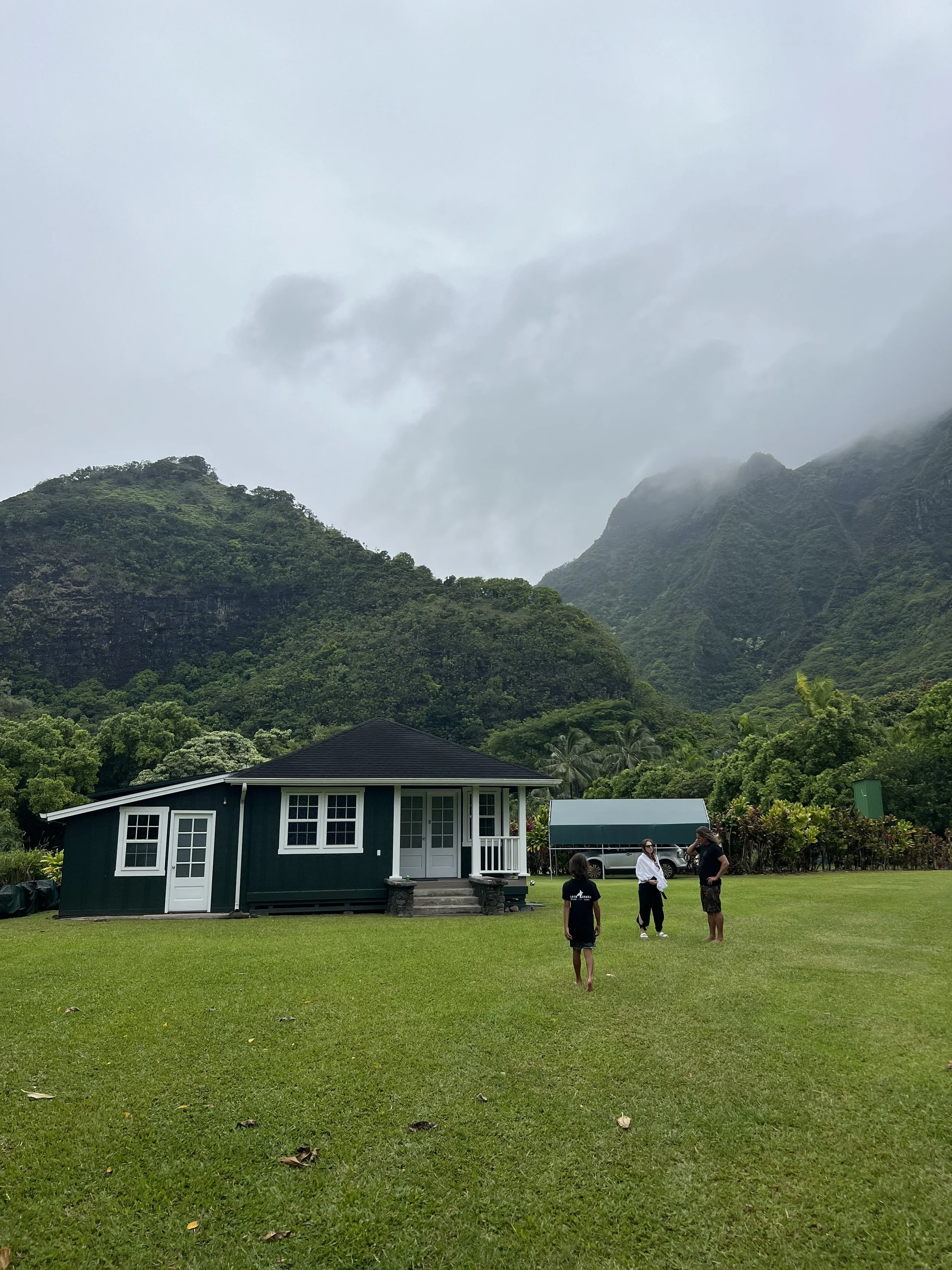 Green house on a grassy field with three people standing nearby, set against lush, green mountains partially covered in clouds.