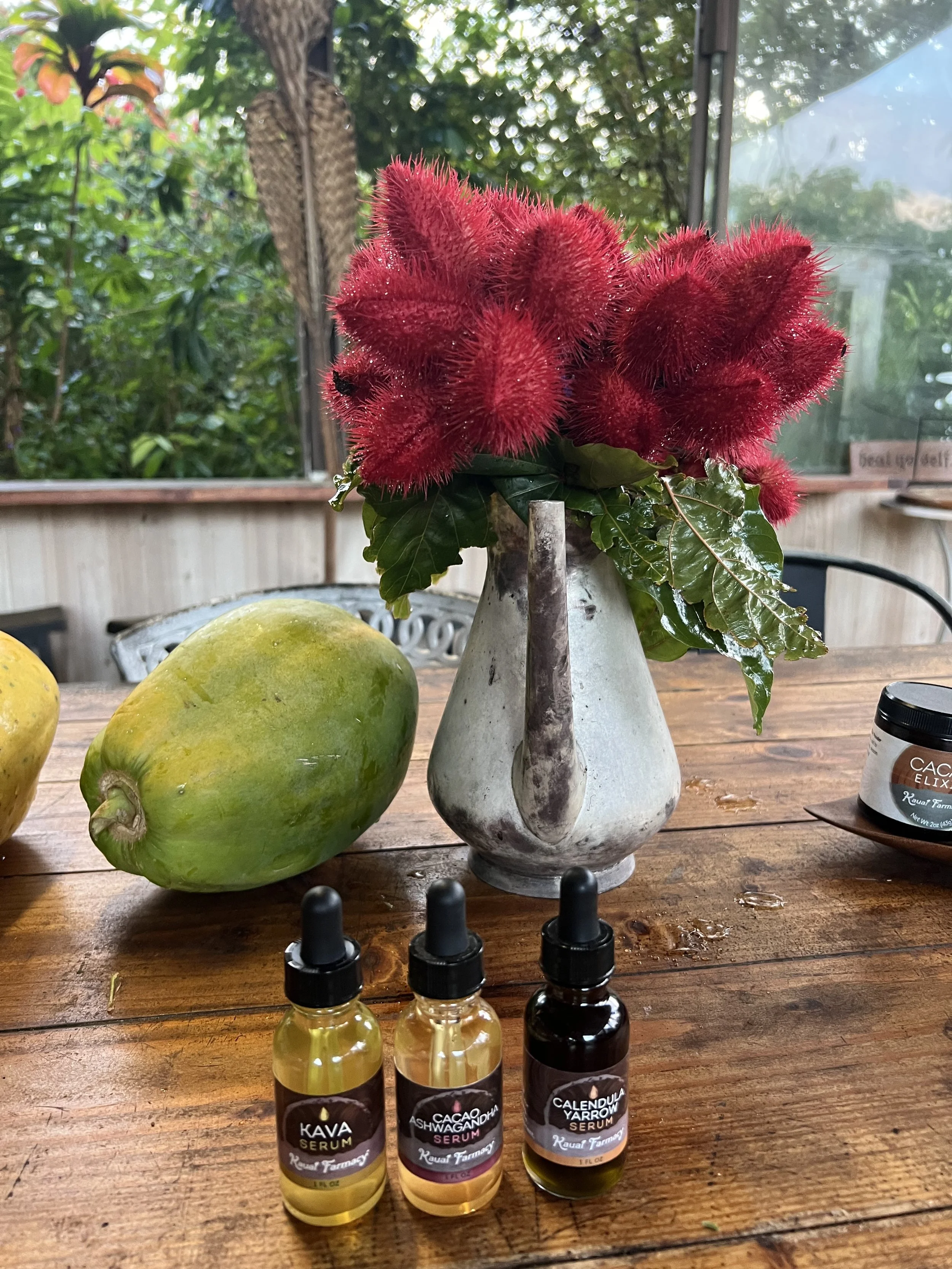 A glass vase with red fuzzy flowers and green leaves, on a wooden table with two papayas, three small bottles of facial serums, and a jar of skincare cream.