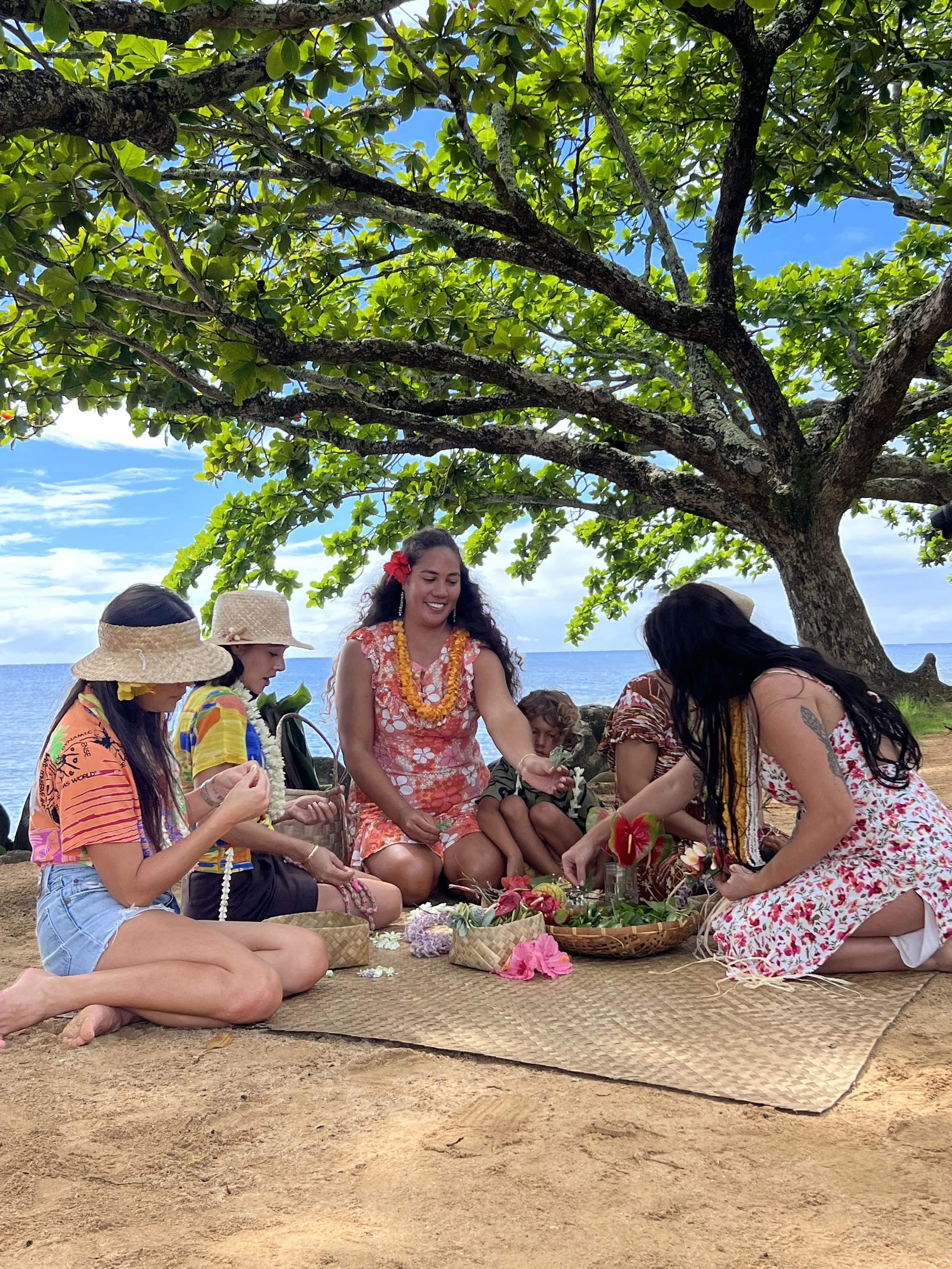 A group of people participating in a traditional Hawaiian ceremony under a large tree on a beach. They are sitting on a woven mat with flowers and ceremonial items, dressed in colorful tropical clothing, with the ocean in the background.