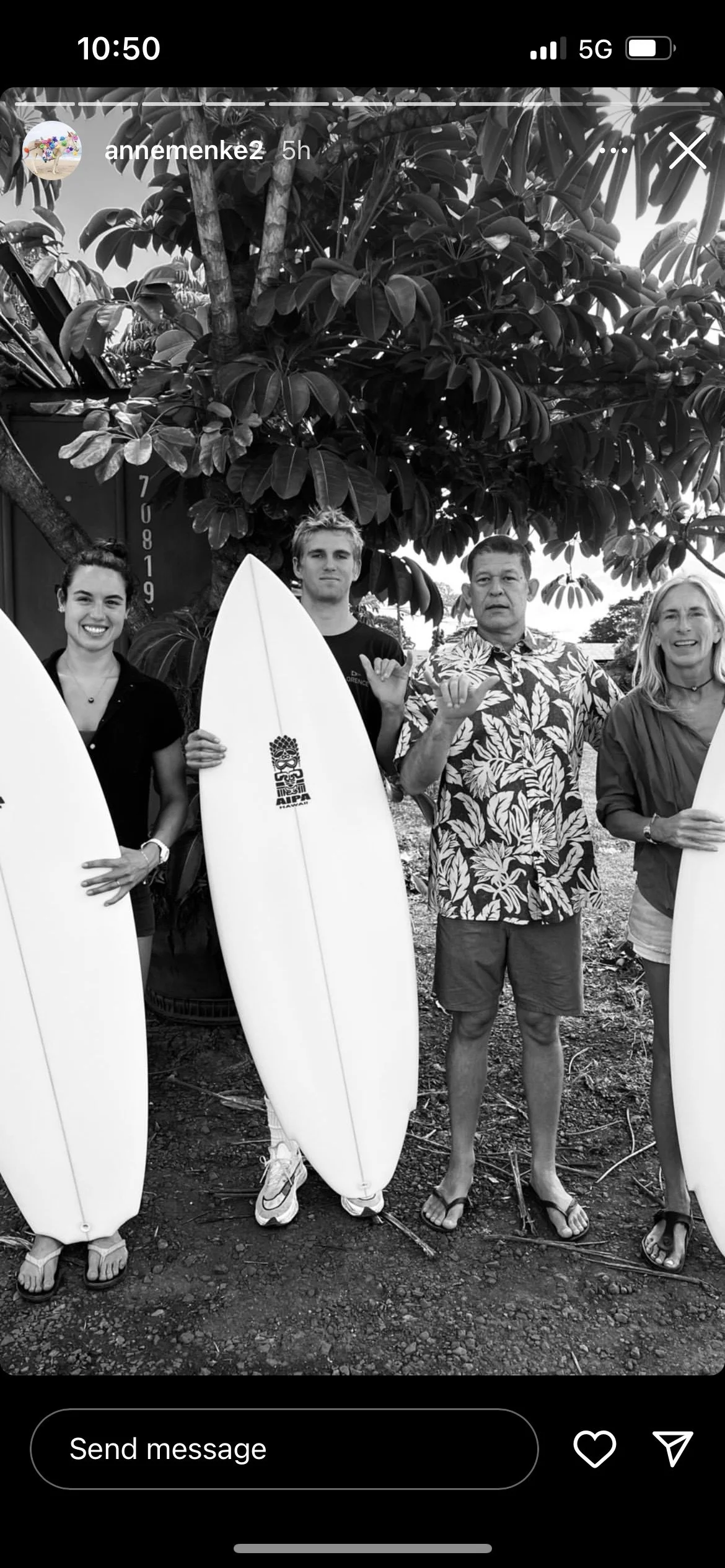 Black and white photo of four people standing outdoors, holding surfboards, with large trees in the background. The people are dressed casually, and one is wearing a Hawaiian shirt.