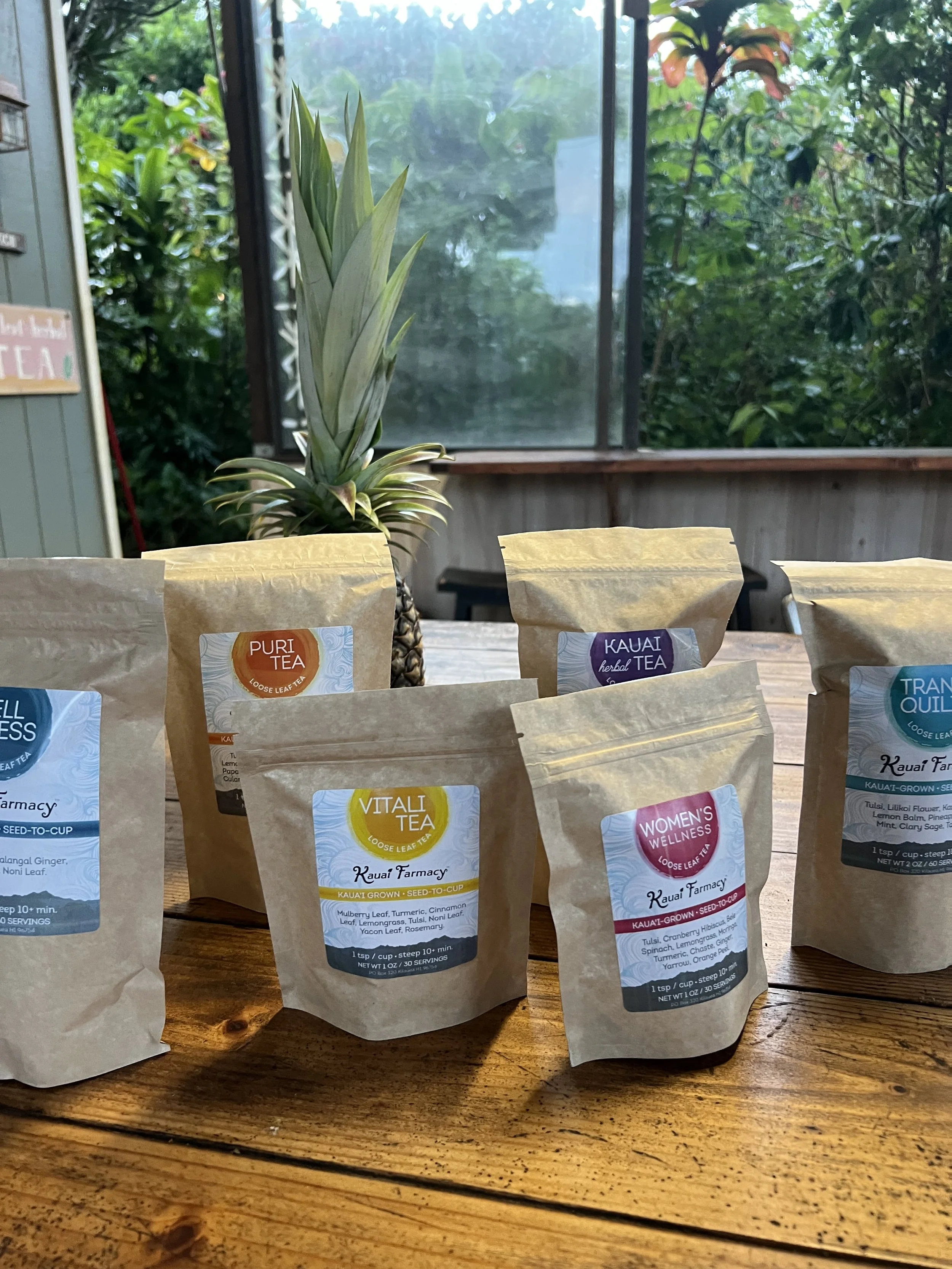 Assorted loose leaf tea bags on a wooden table with a pineapple in the background and greenery outside a screened porch.