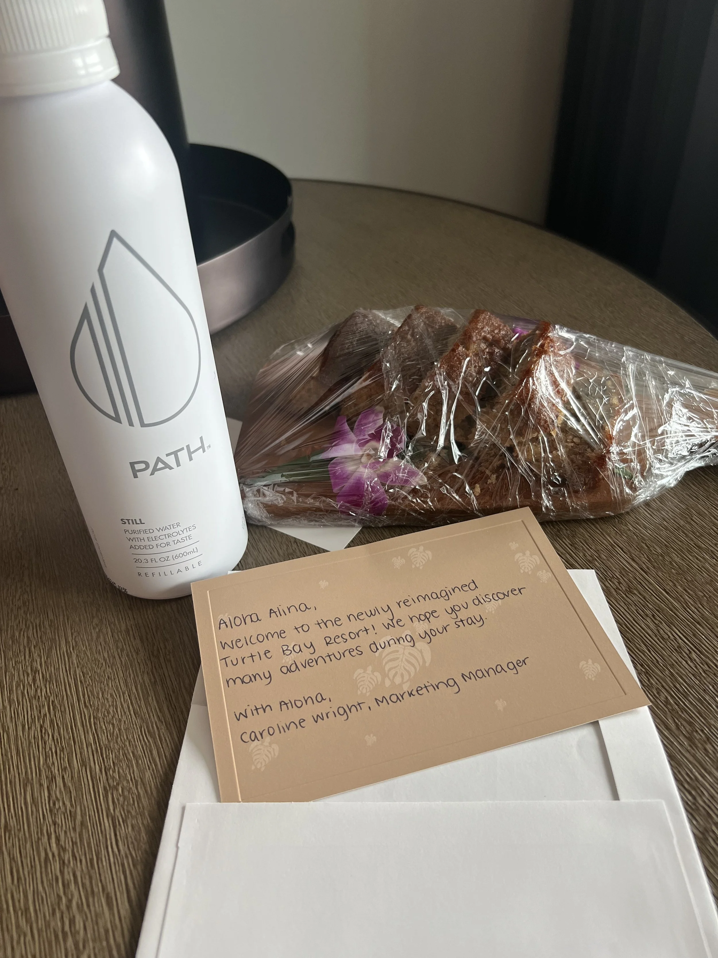 A white bottle of PATH brand bottled water, a clear plastic bag of sliced bread with purple flowers, and a welcome note on the table. The note says, 'Aloha Aina, Welcome to the newly reimagined Turtle Bay Resort! We hope you discover many adventures
