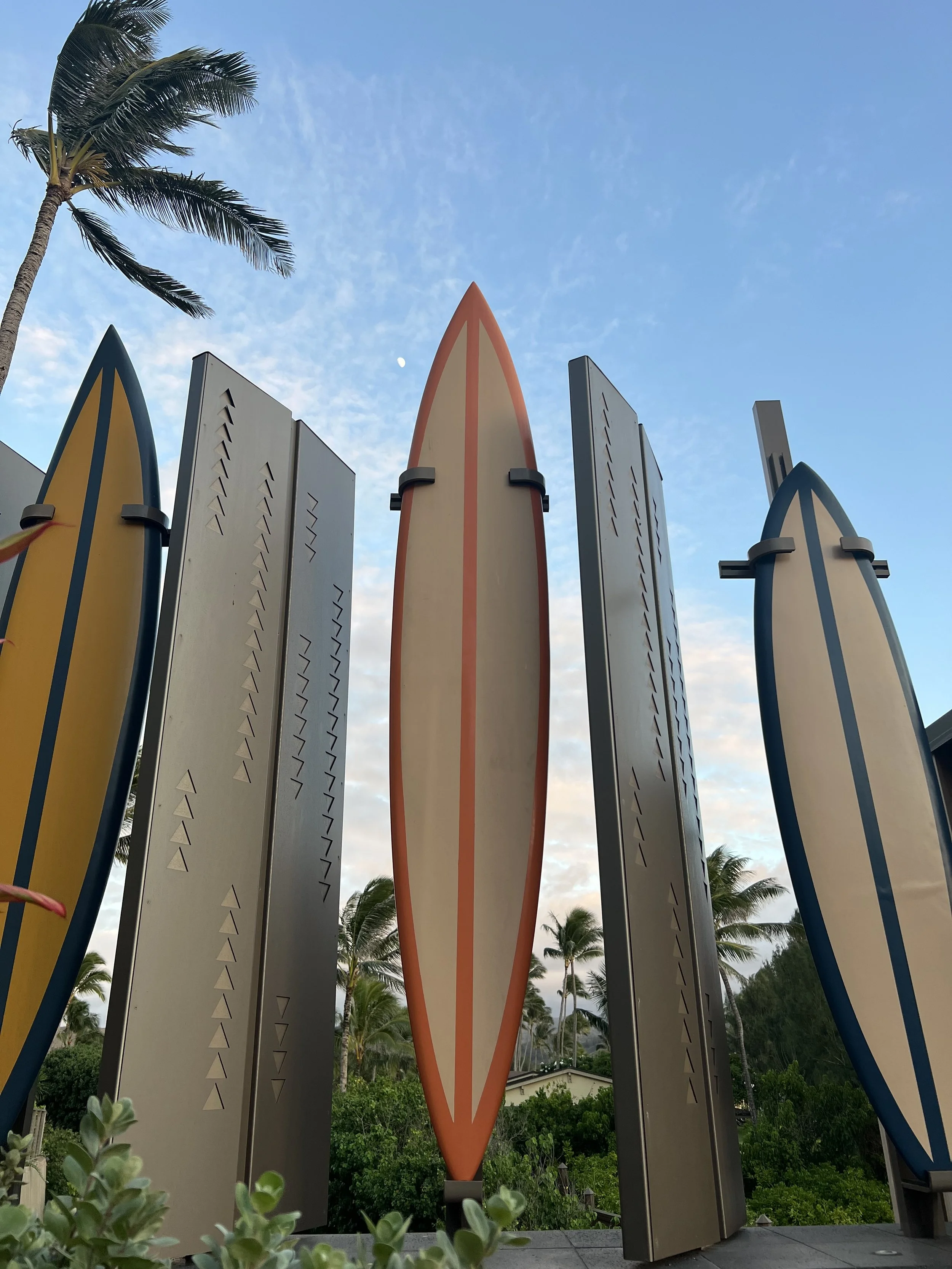 Multiple colorful surfboards are displayed vertically outdoors with palm trees and a blue sky in the background.