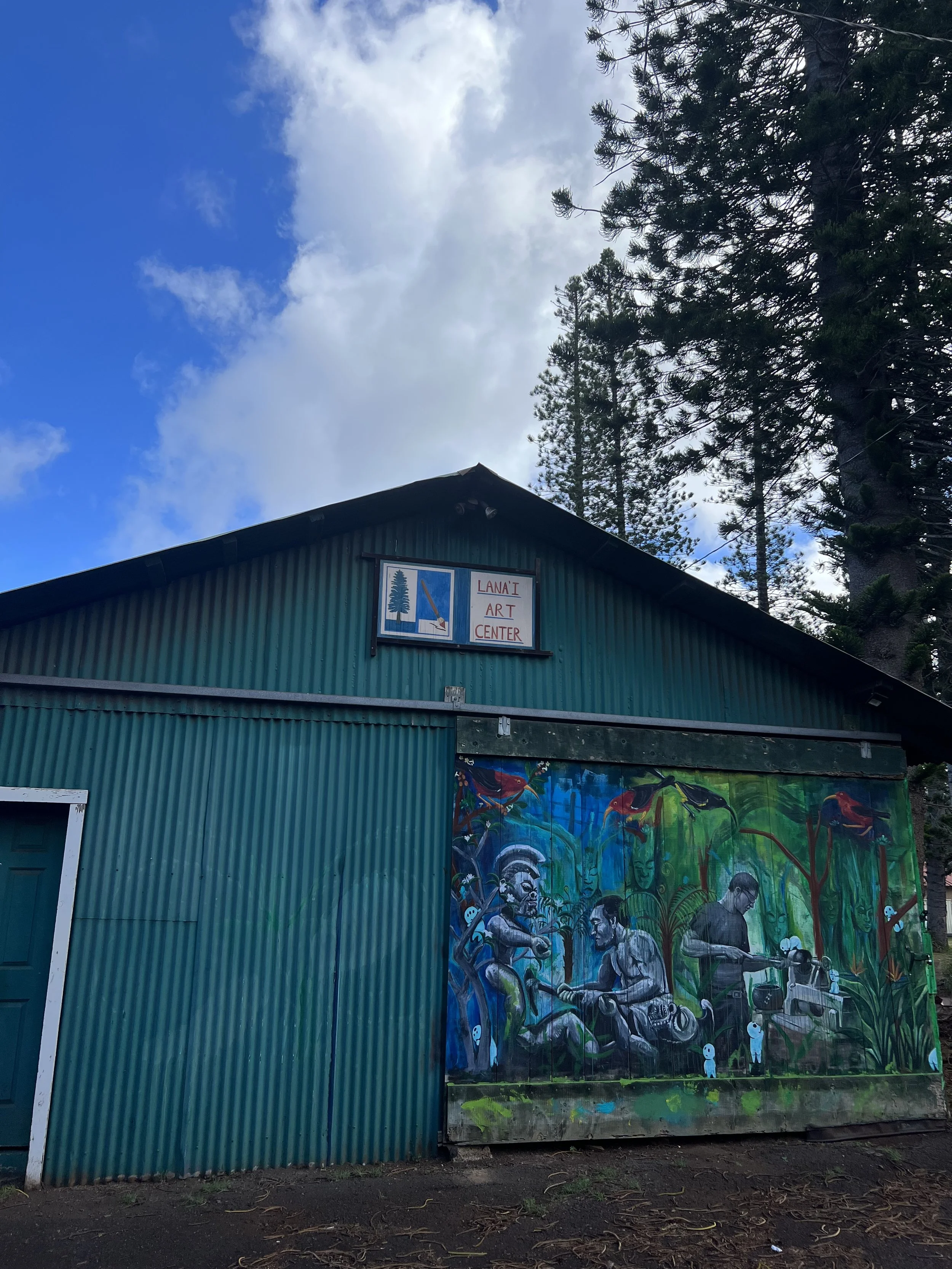 A green building with a sign reading 'Lana'i Art Center' on the upper part of the structure, with a colorful mural featuring people and nature scenes painted on the lower part. Tall trees are visible in the background under a partly cloudy sky.