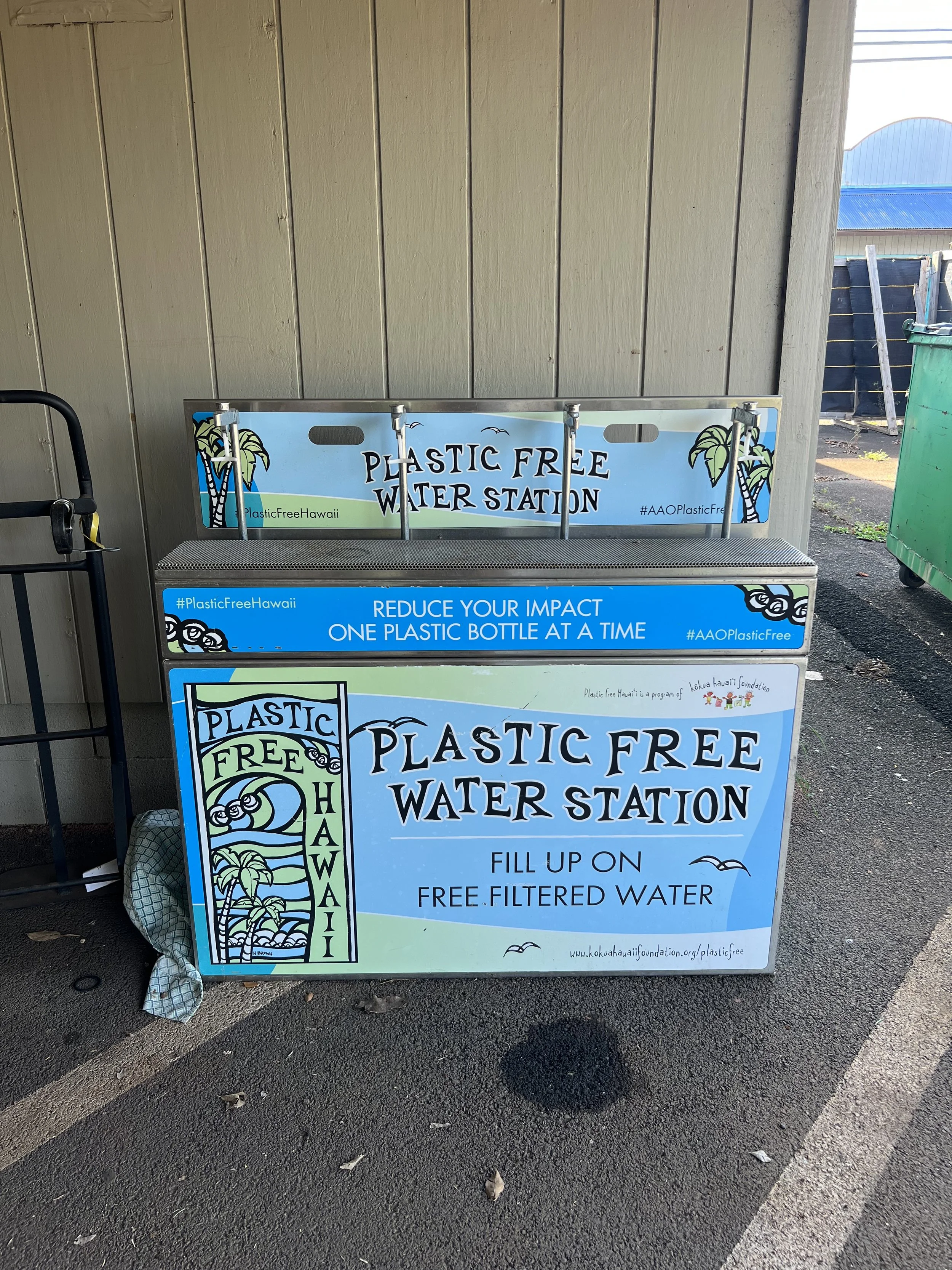 A sign for a plastic-free water station encouraging filling bottles with free filtered water, located near a beige wall and a parking lot.