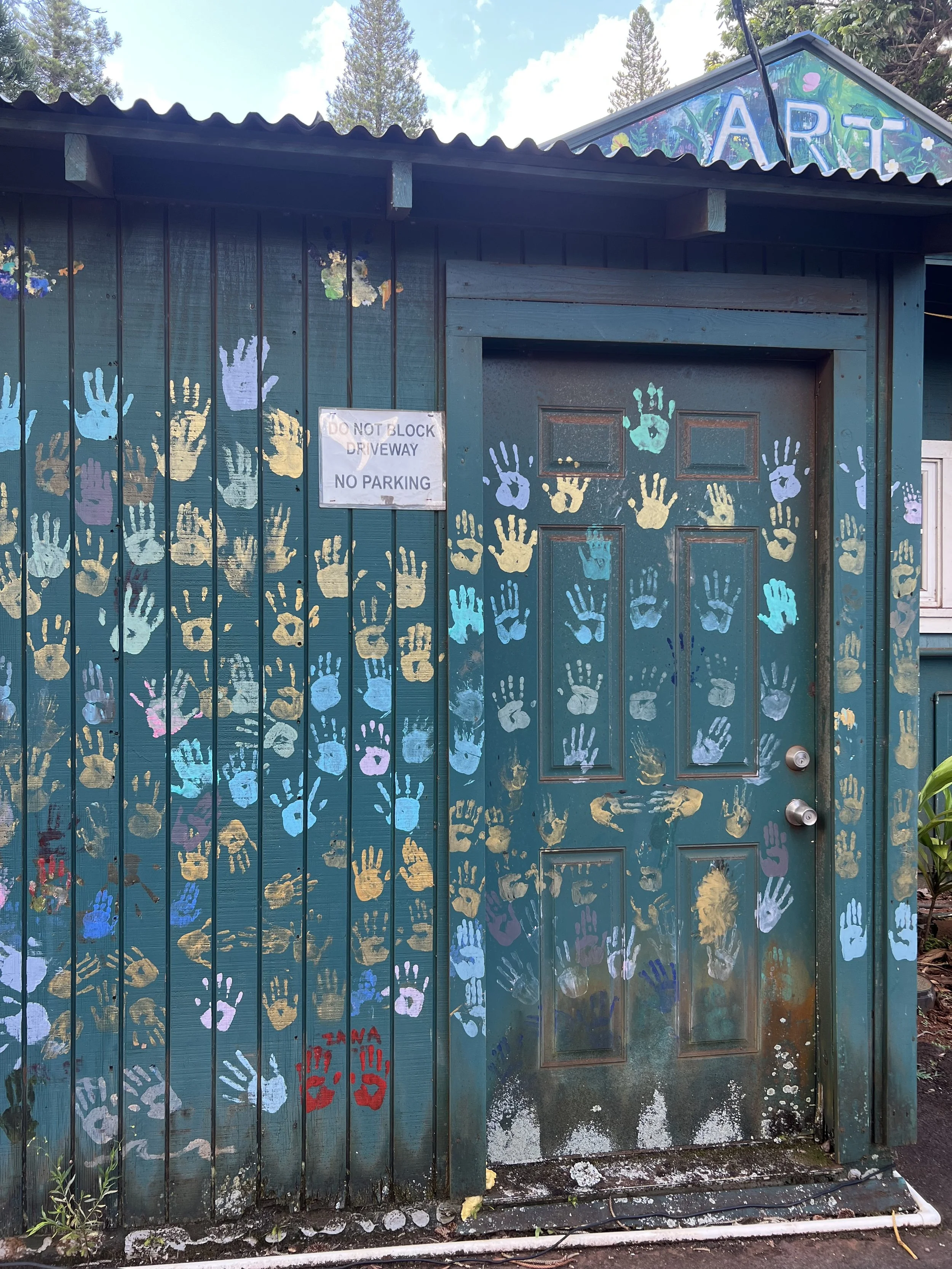 A teal wooden building with a door and siding decorated with colorful handprints in shades of yellow, blue, purple, and pink. A sign on the siding reads 'DO NOT BLOCK DRIVEWAY NO PARKING.' The top of the building has a painted mural with the letters
