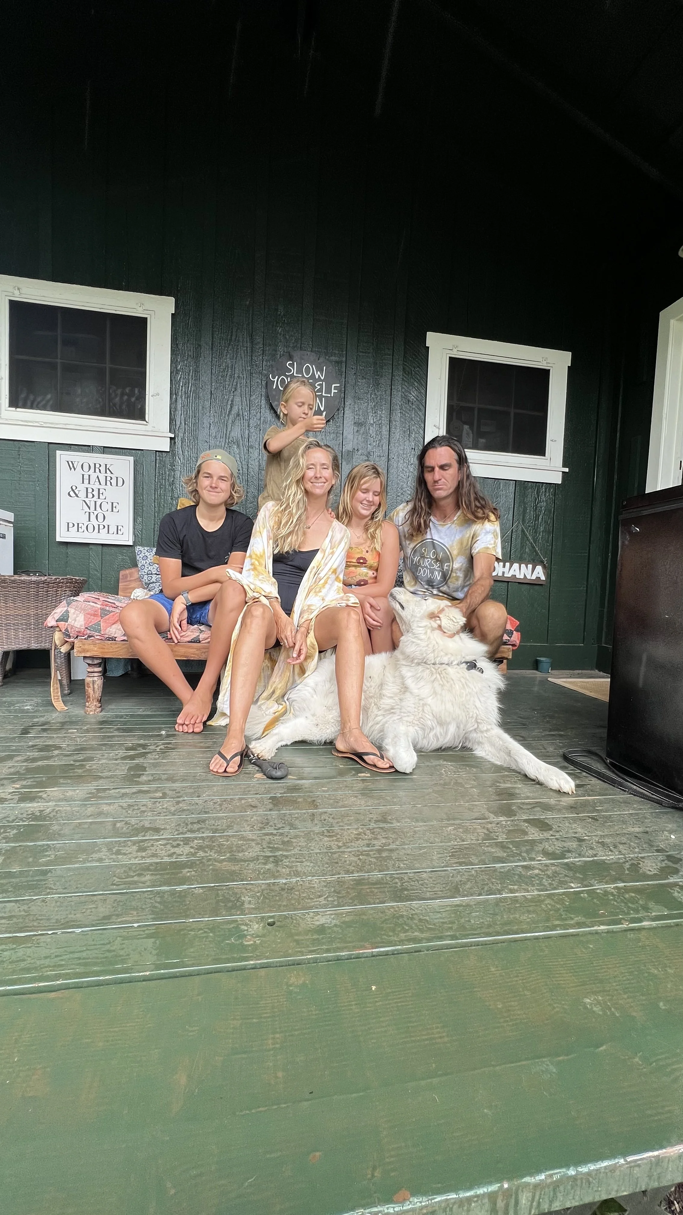 A family of five, including children and a man, sitting on a porch with a large white dog. The porch has a green wooden floor and dark green walls with white trim, and two signs that read 'Work Hard & Be Nice to People' and 'Slow Your Self Down'.
