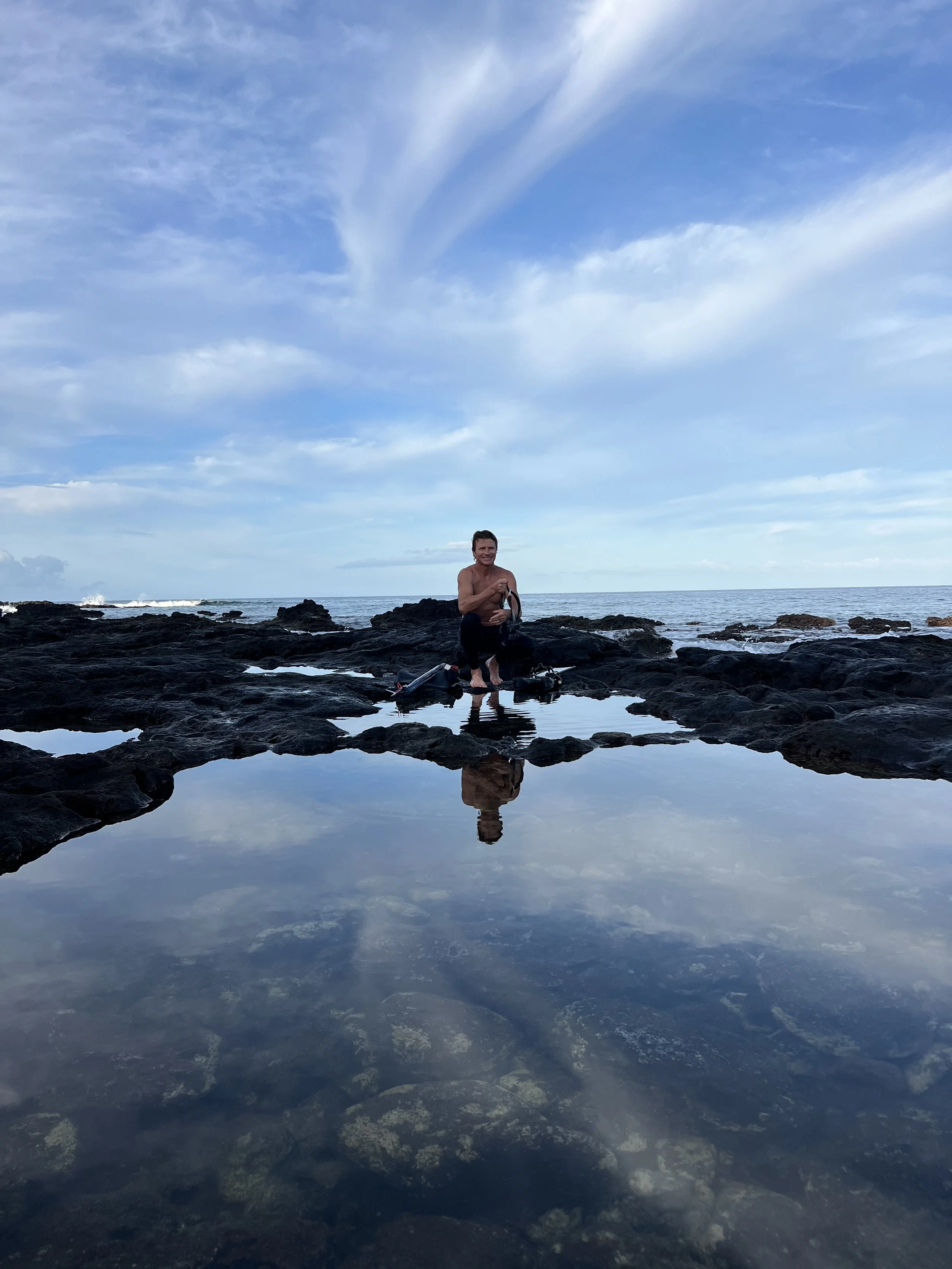 A man kneeling on dark rocks at the shoreline with the ocean and a cloudy sky in the background, taking a photo with his phone, along with a reflection of him and the sky in a tide pool.