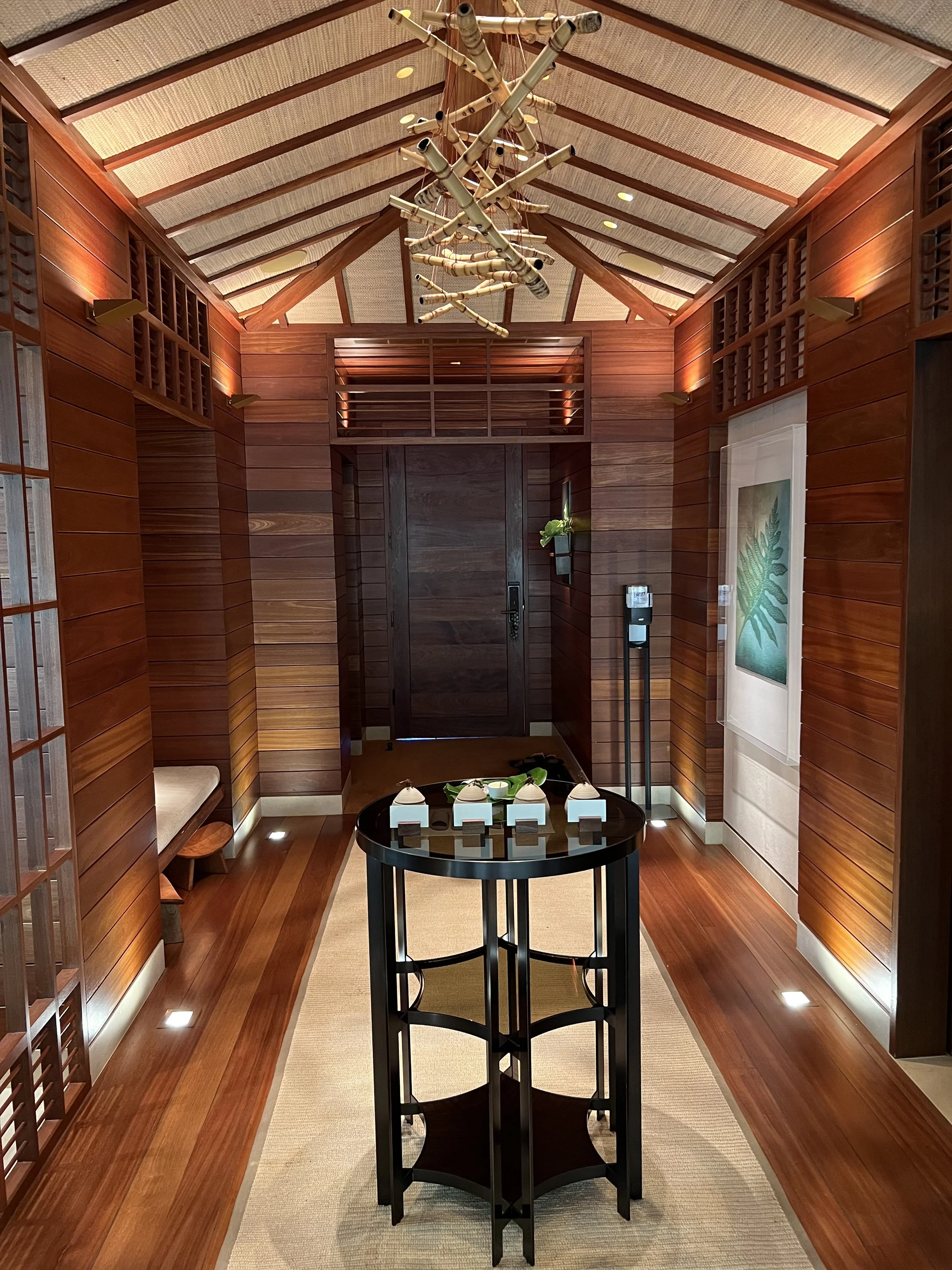 Interior of a modern, wood-paneled hallway with a high, vaulted ceiling, a chandelier hanging from the ceiling, a black round table with small ceramic items on it, a bench on the left, and a framed artwork on the right wall.