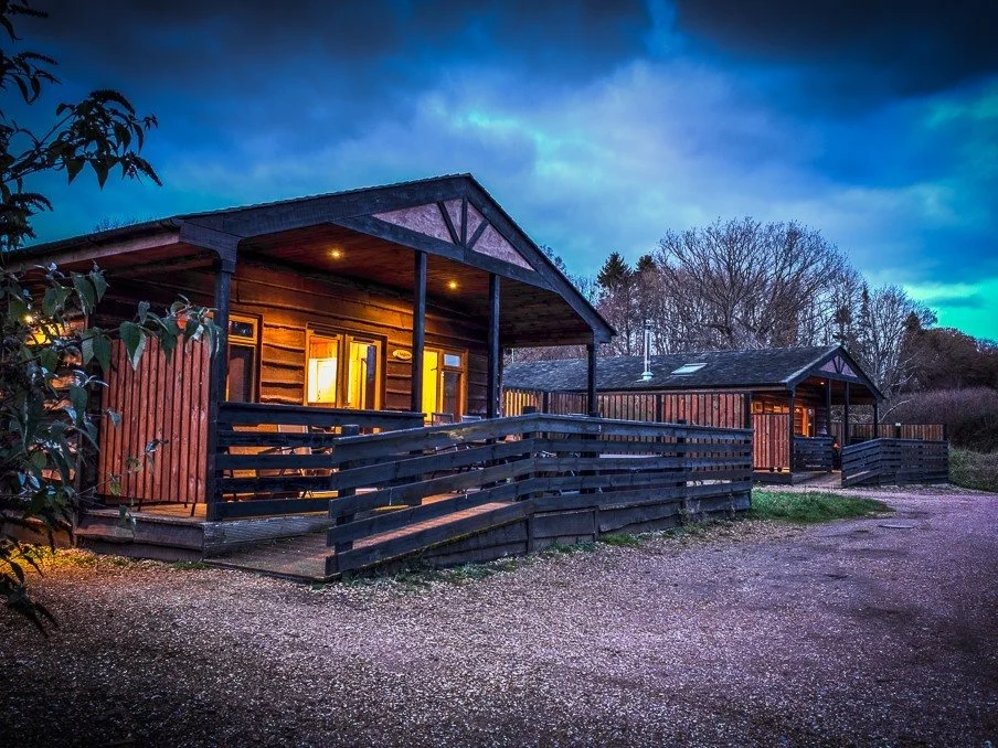 Two wooden cabins with porch railings, illuminated windows, and dark roofs, are set against a backdrop of trees under a mostly cloudy sky at dusk.