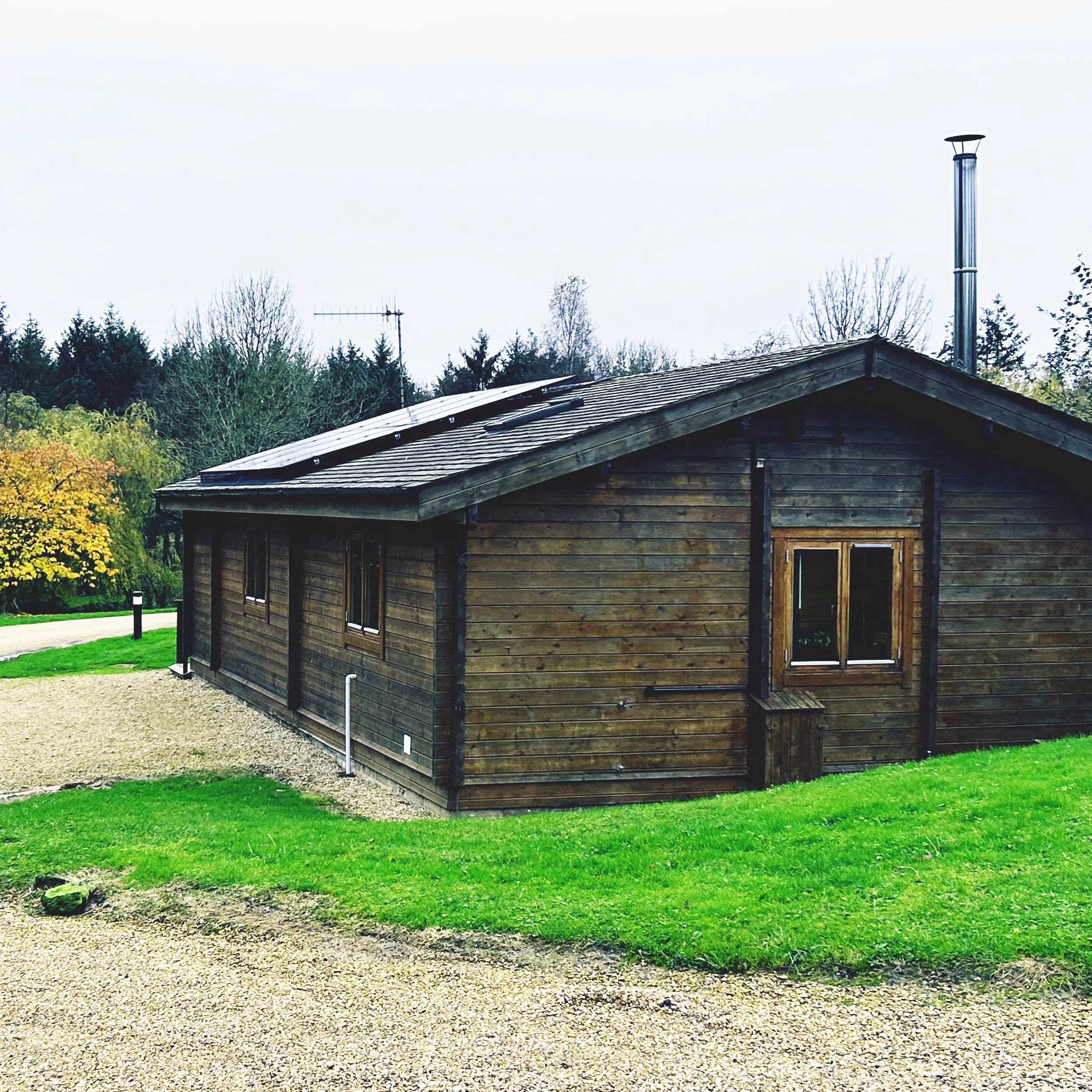 A wooden house with solar panels on the roof, surrounded by greenery and trees in the background.