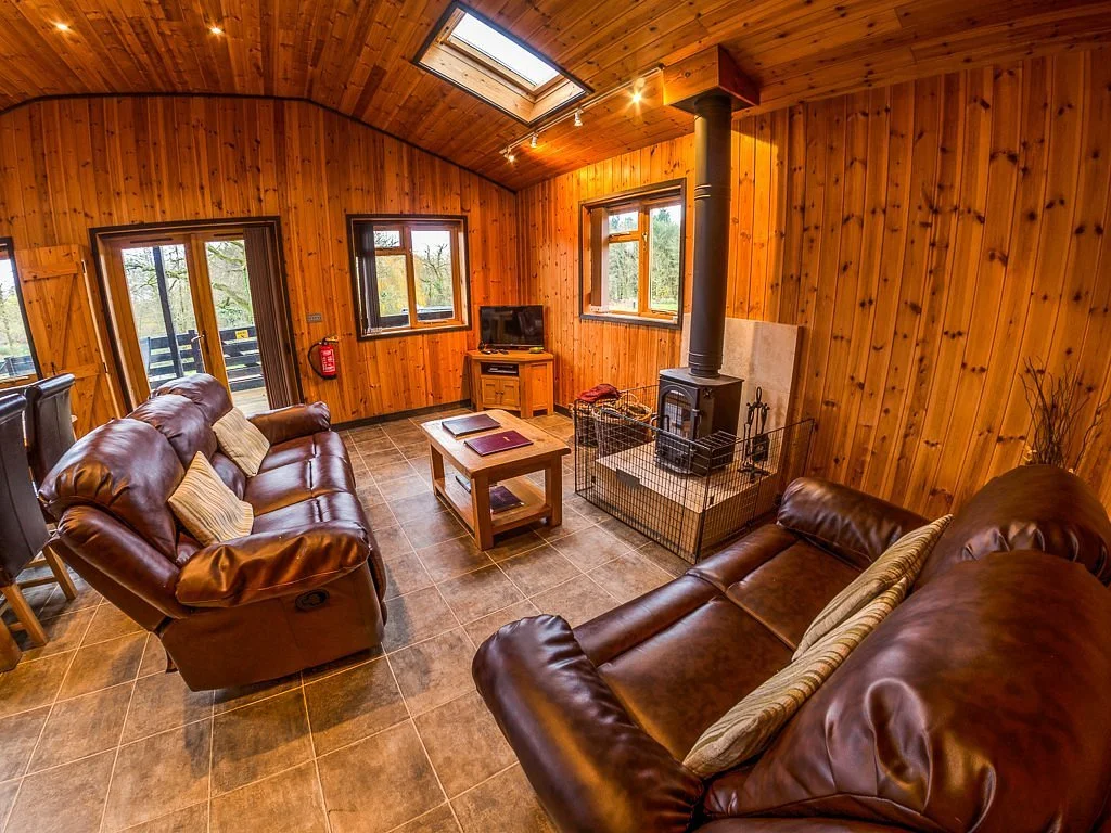 Cozy living room with wooden walls and ceiling, featuring two leather sofas, a central wooden coffee table, a small television on a wooden stand, and a black wood-burning stove with a metal fence around it. There are three windows and a glass door leading outside, along with a skylight letting in natural light.