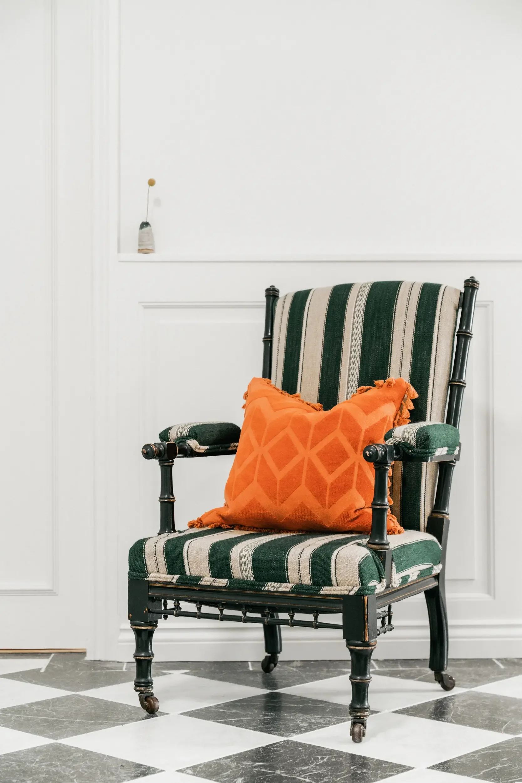 A vintage style chair with black frame and green and beige striped upholstery, featuring a vibrant orange pillow with a geometric pattern, set against a white paneled wall and black and white checkered floor.